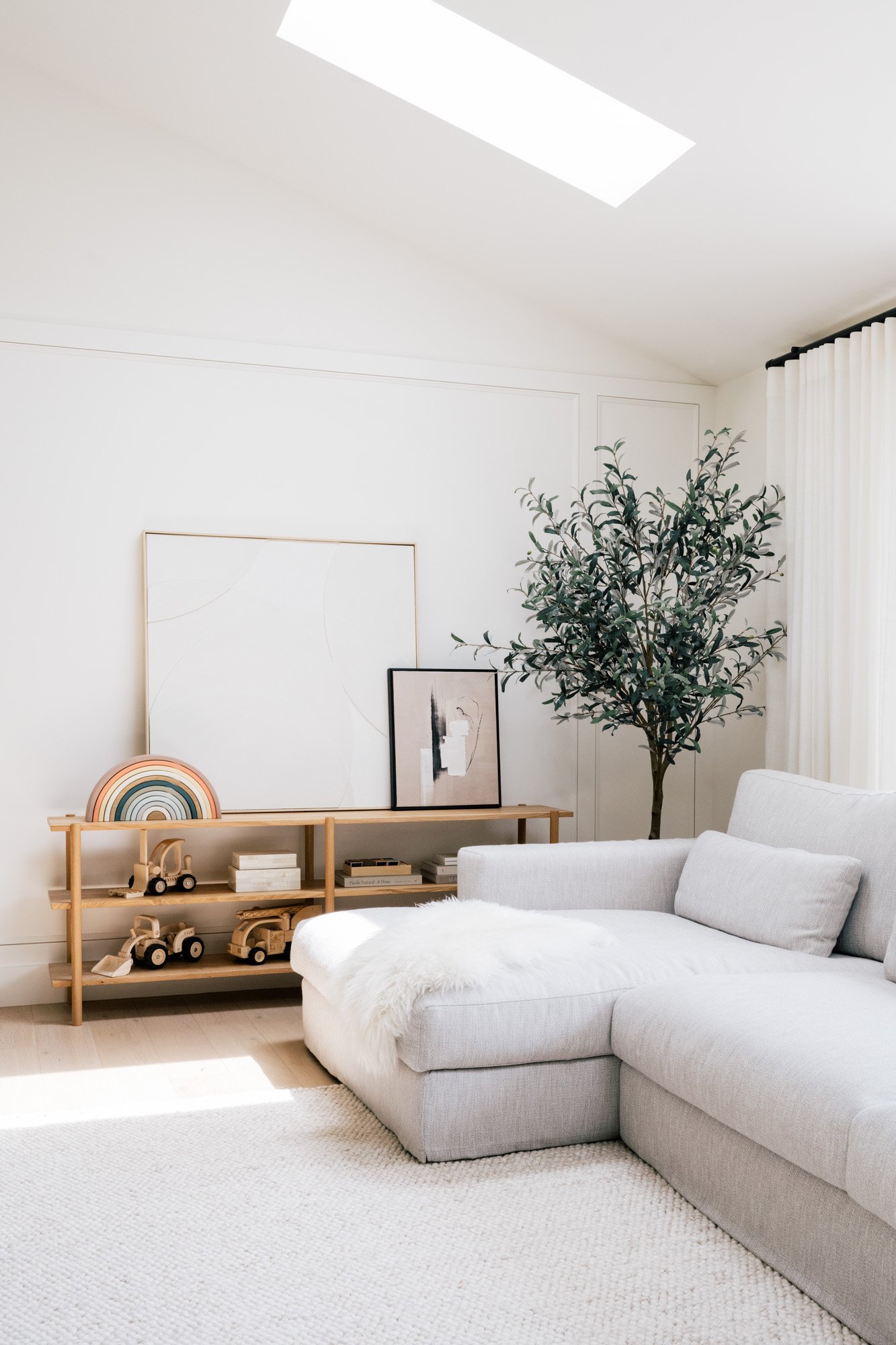 A large light 3-seater grey couch sits in front of a bay window with a Kroft shelf next to it. Sunlight comes in from the window and the vaulted ceiling's skylight