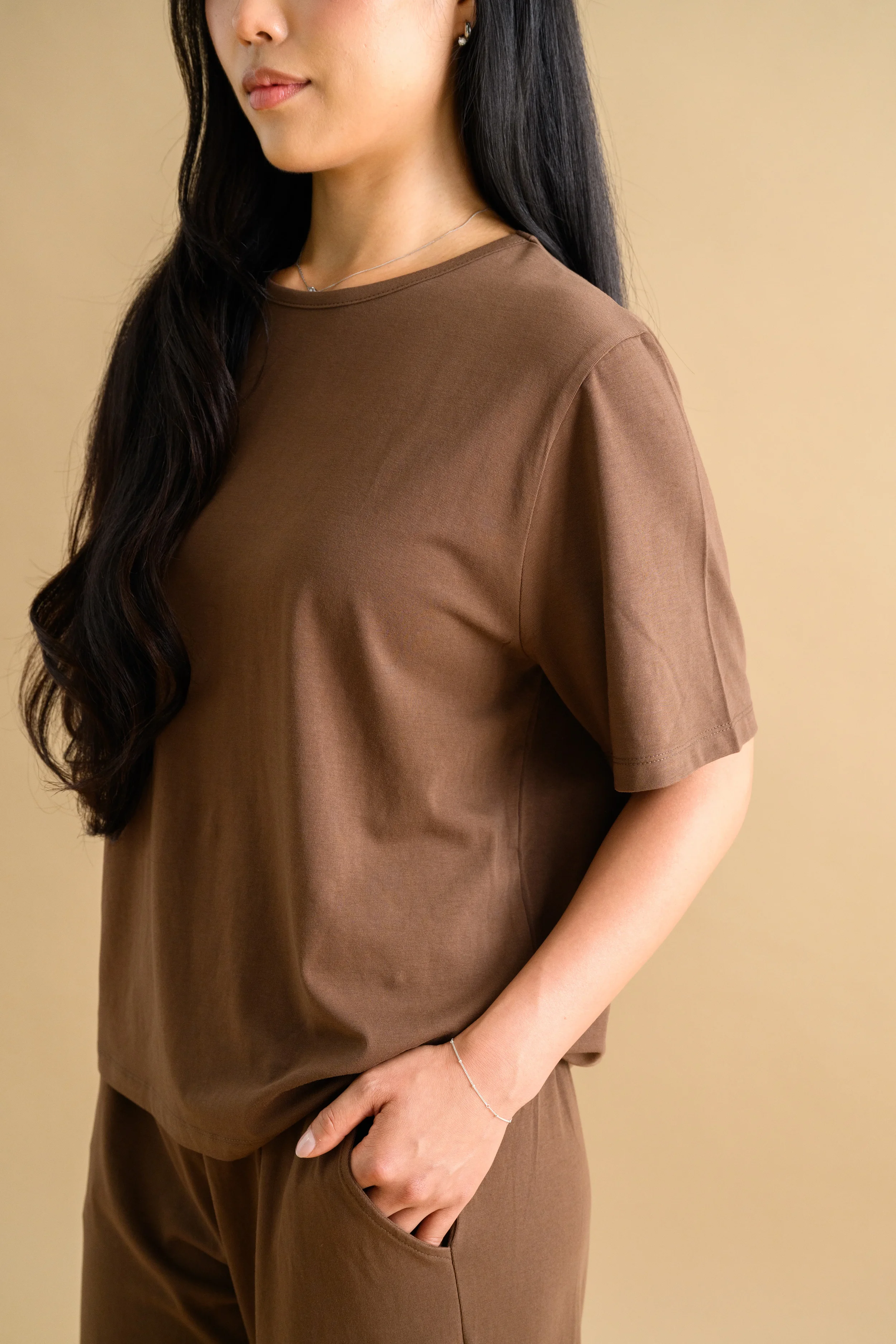 A woman standing against a beige backdrop, wearing a brown short sleeved shirt and long loose pants by Heirloome House.