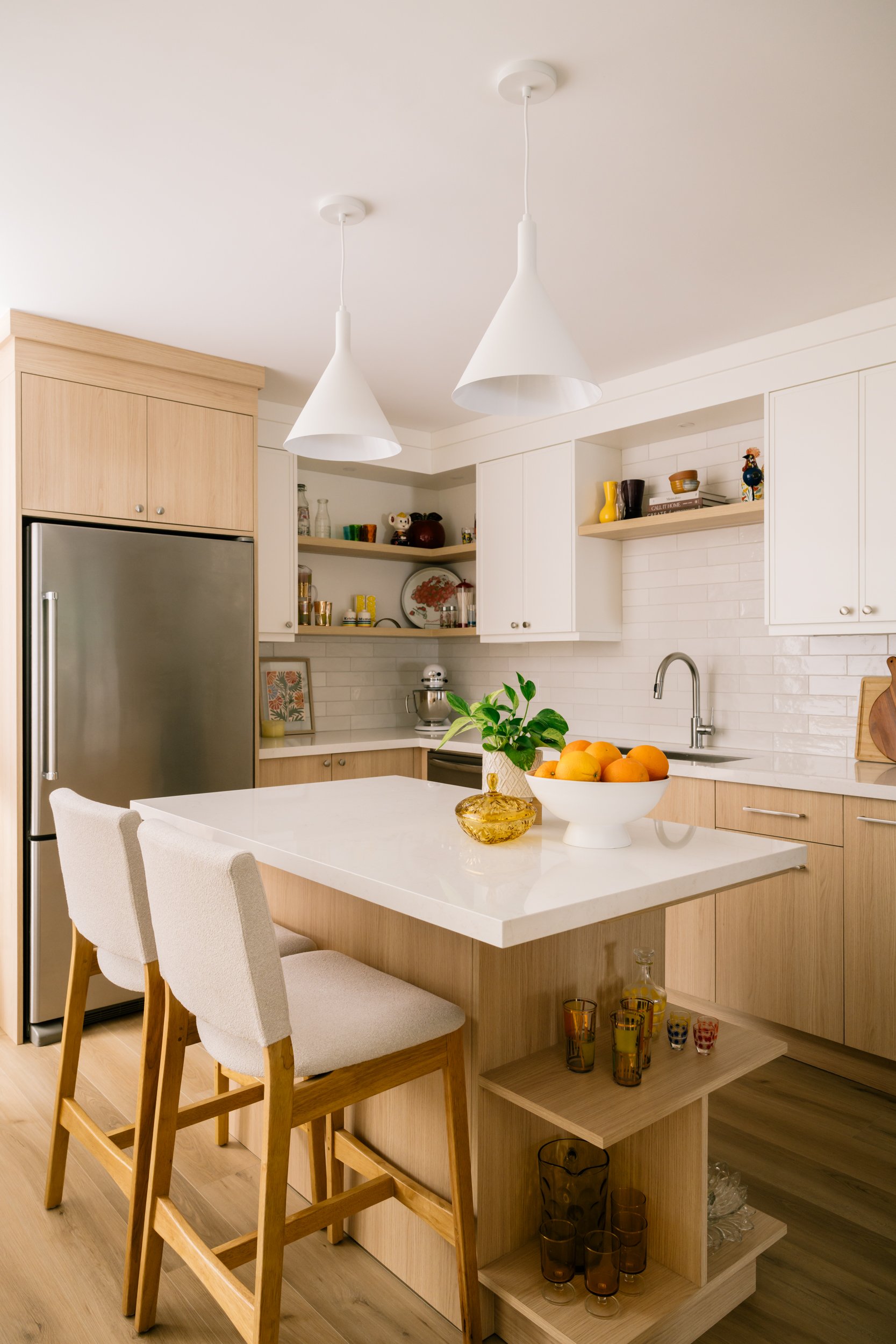 A kitchen with white upper cabinets and light wood lower cabinets. A small kitchen island in the middle of the kitchen with two stools and two pendants lights above.