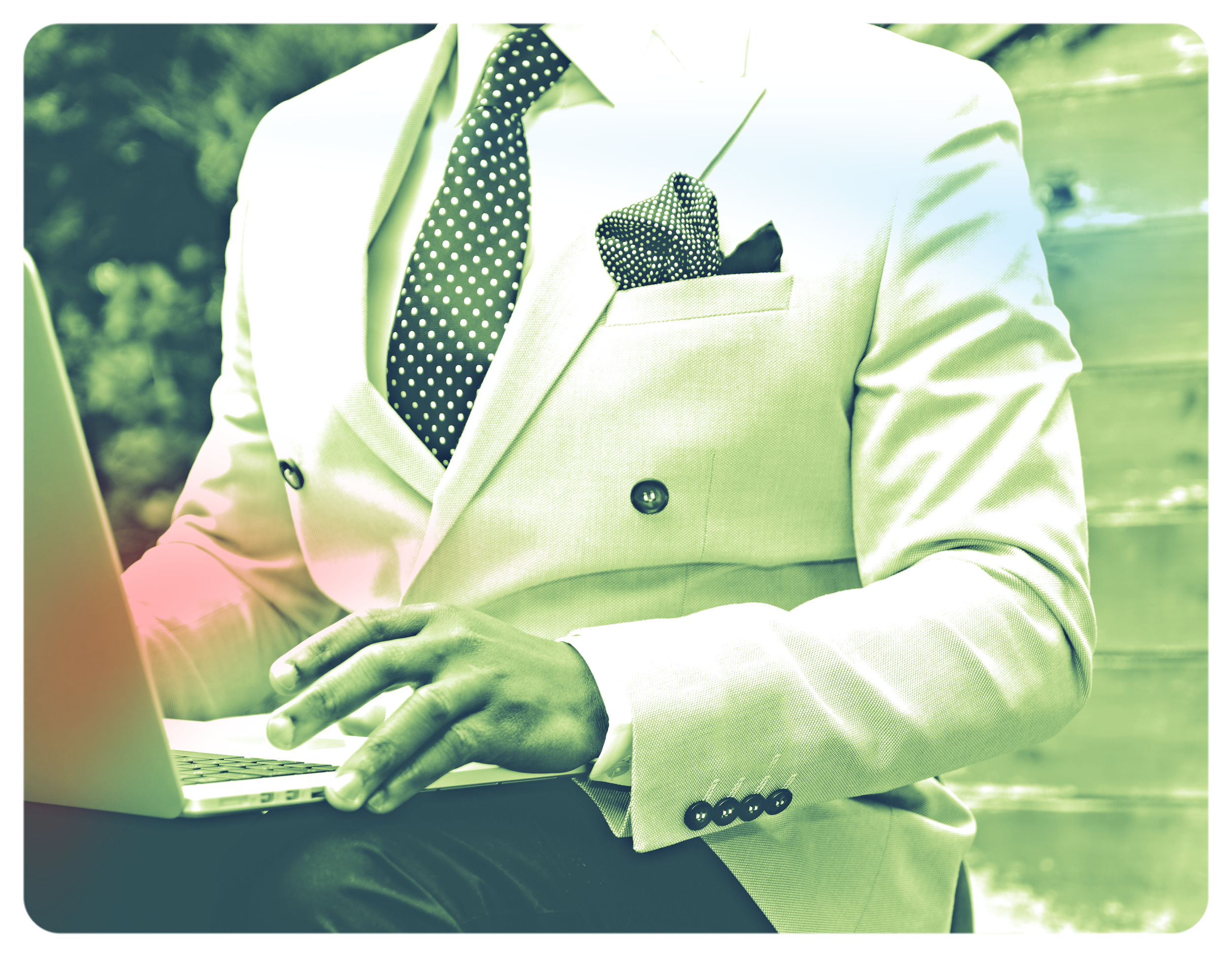 A man dressed in a light-colored suit with a polka dot tie and pocket square, sitting outdoors and using a laptop.