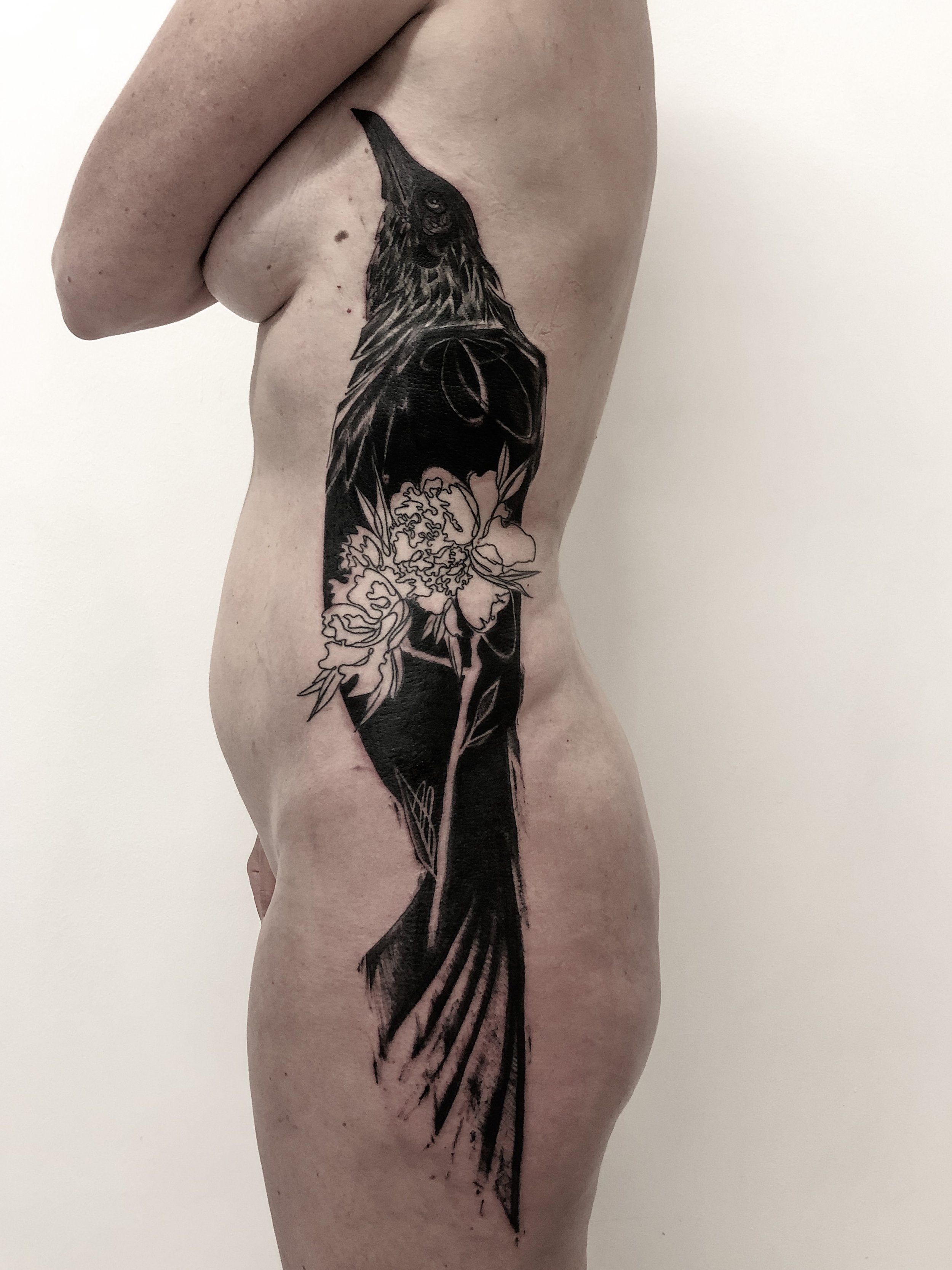 Tatouage Réaliste de Corbeau Noir Tenant une Fleur Blanche sur le Côté du Corps”