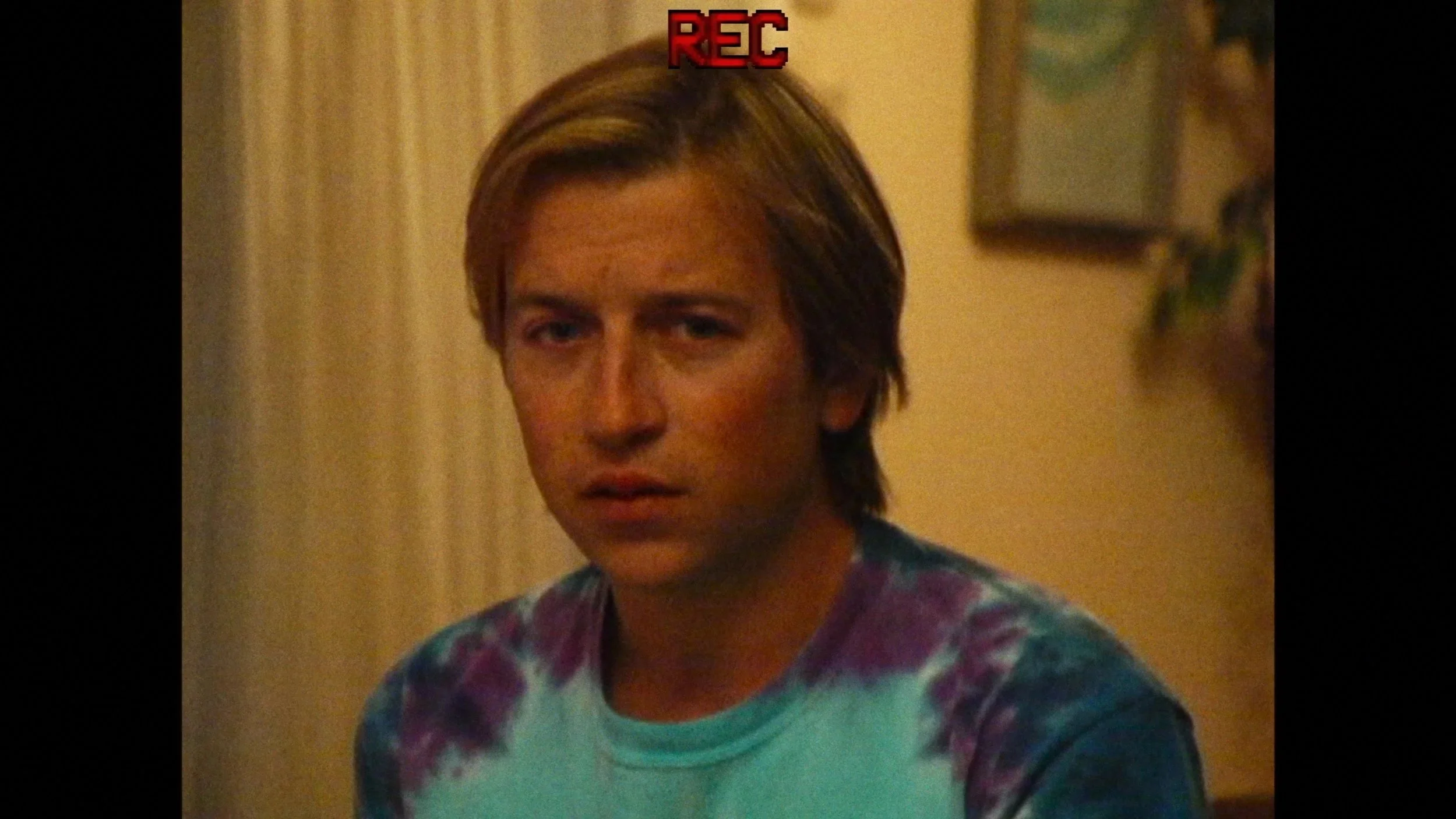 Actor Skyler Gisondo wears a tye-died purple and blue t-shirt and stares at the camera with a concerned look on his face. The image is grainy, as if viewed through a recording camera
