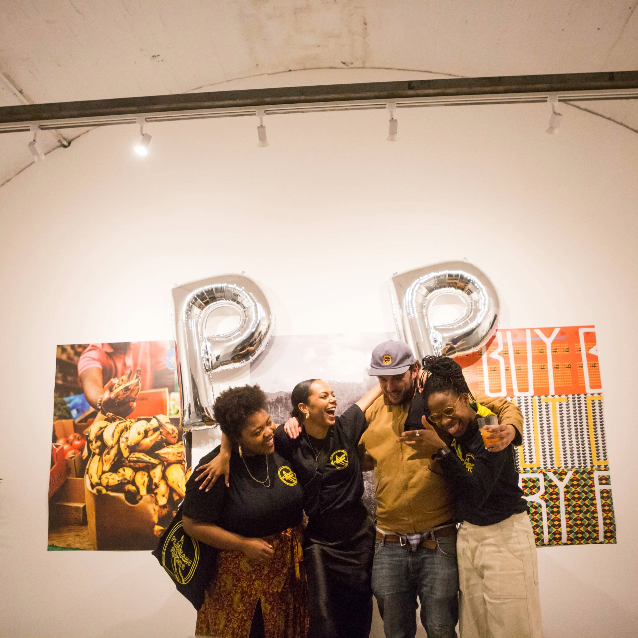 Four people smiling and celebrating at an event with large silver balloons forming the letters P and P in the background. They are standing in front of a colorful patterned backdrop and wall art.