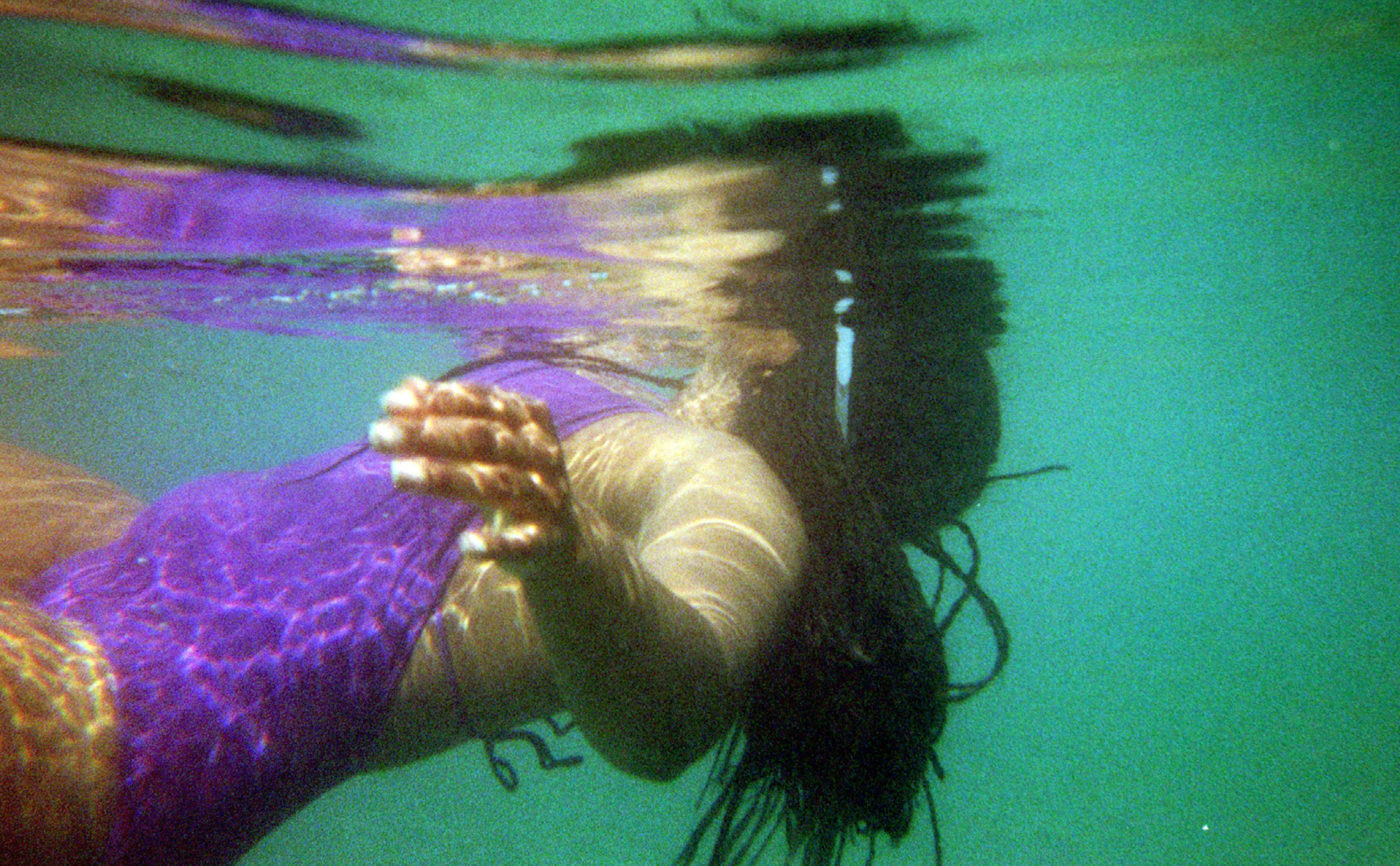 A woman underwater wearing a purple swimsuit, with her hair flowing, and her right hand on the back of her neck.