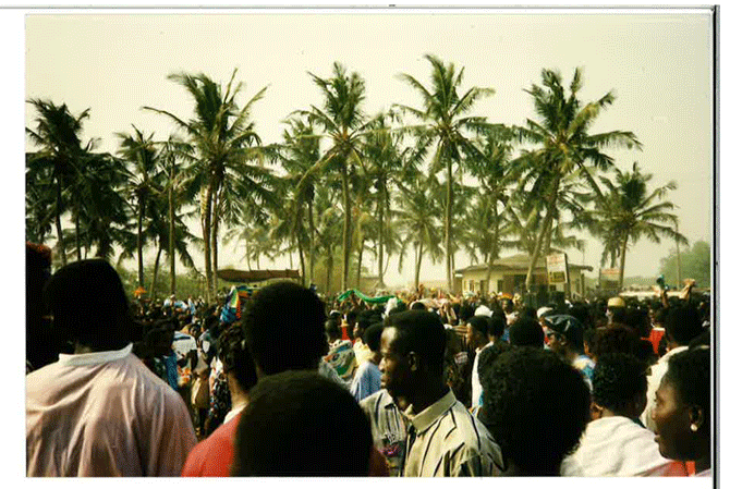People gathered outdoors among tall palm trees, with some structures and vehicles visible in the background.