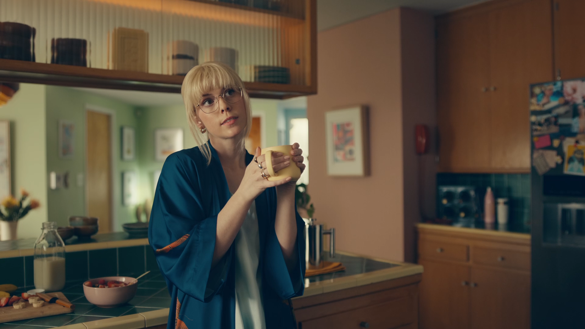 A young woman with blonde hair and glasses holding a yellow mug in a kitchen with green walls and wooden cabinets, surrounded by various breakfast items and fruit on the counter.