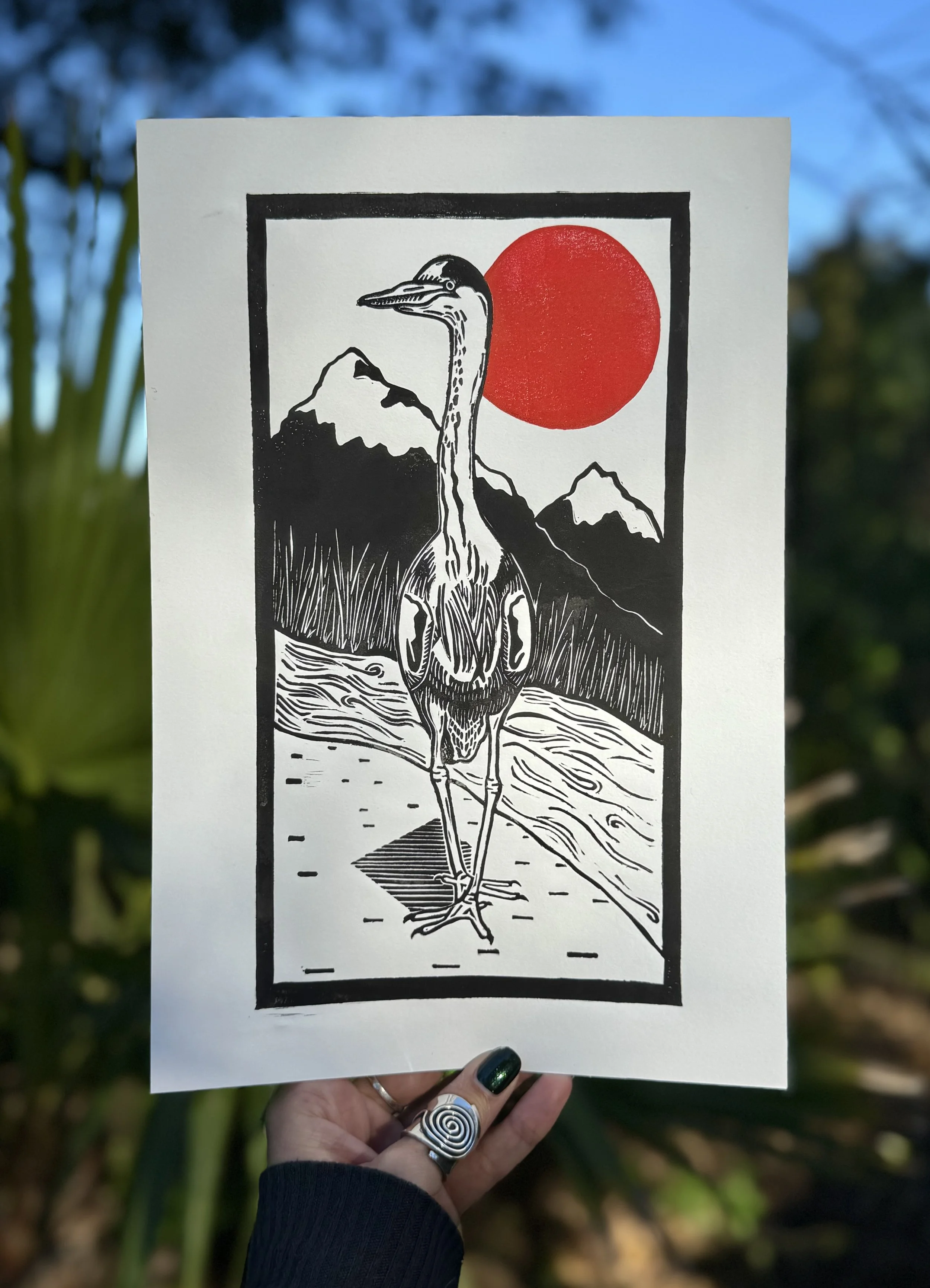 A black and red linoprint of a heron standing in front of a river, reeds, and mountains with a red sun hanging in the sky