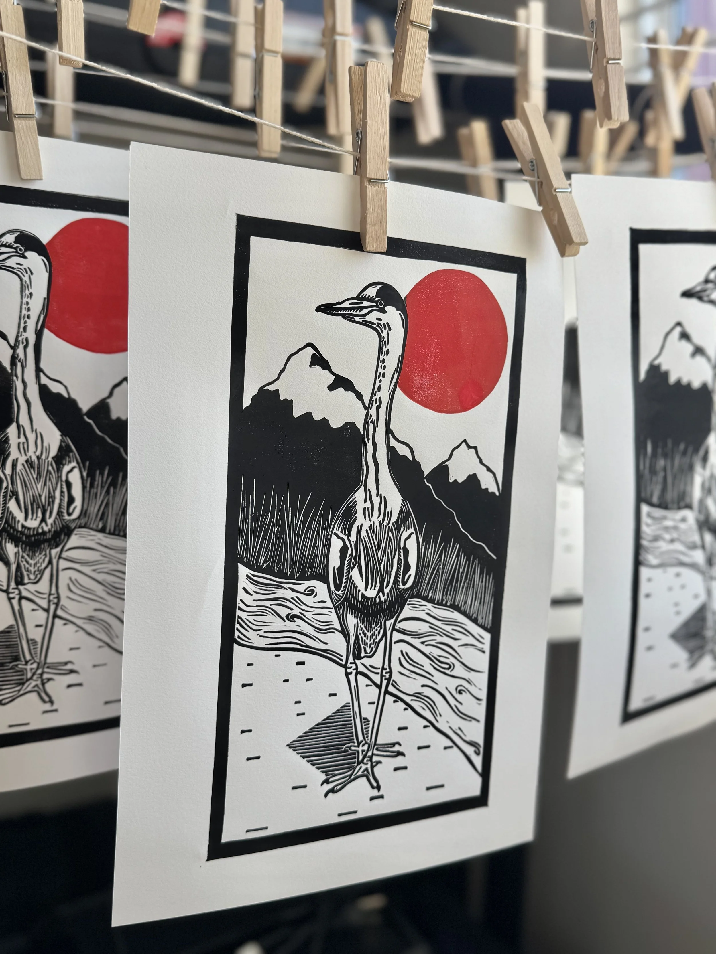 A black and red linoprint of a heron standing in front of a river, reeds, and mountains with a red sun hanging in the sky. Three linoprints hang from clothespins on a drying rack