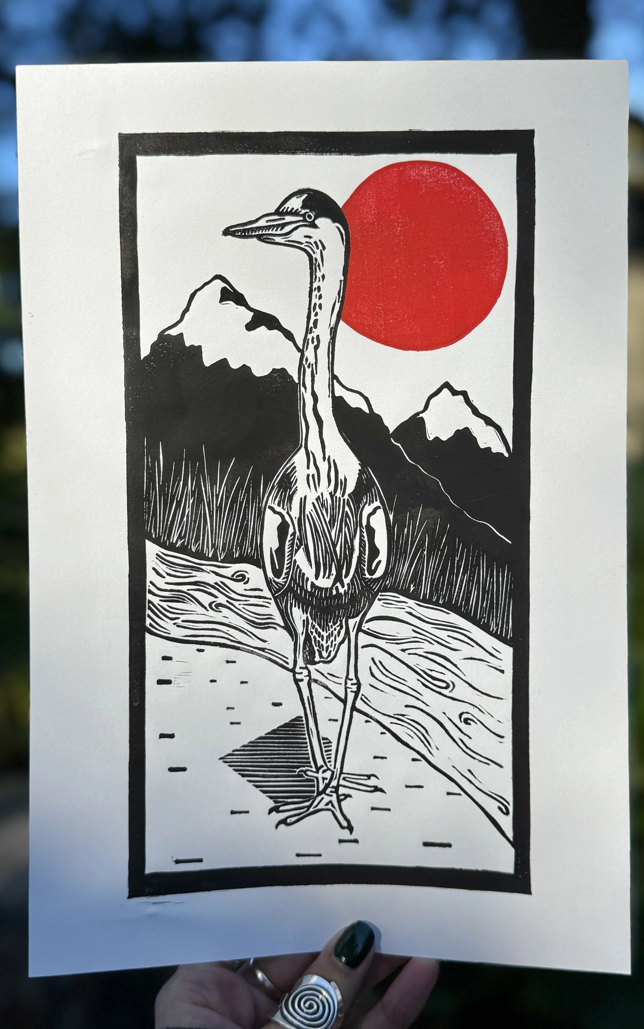 A black and red linoprint of a heron standing in front of a river, reeds, and mountains with a red sun hanging in the sky