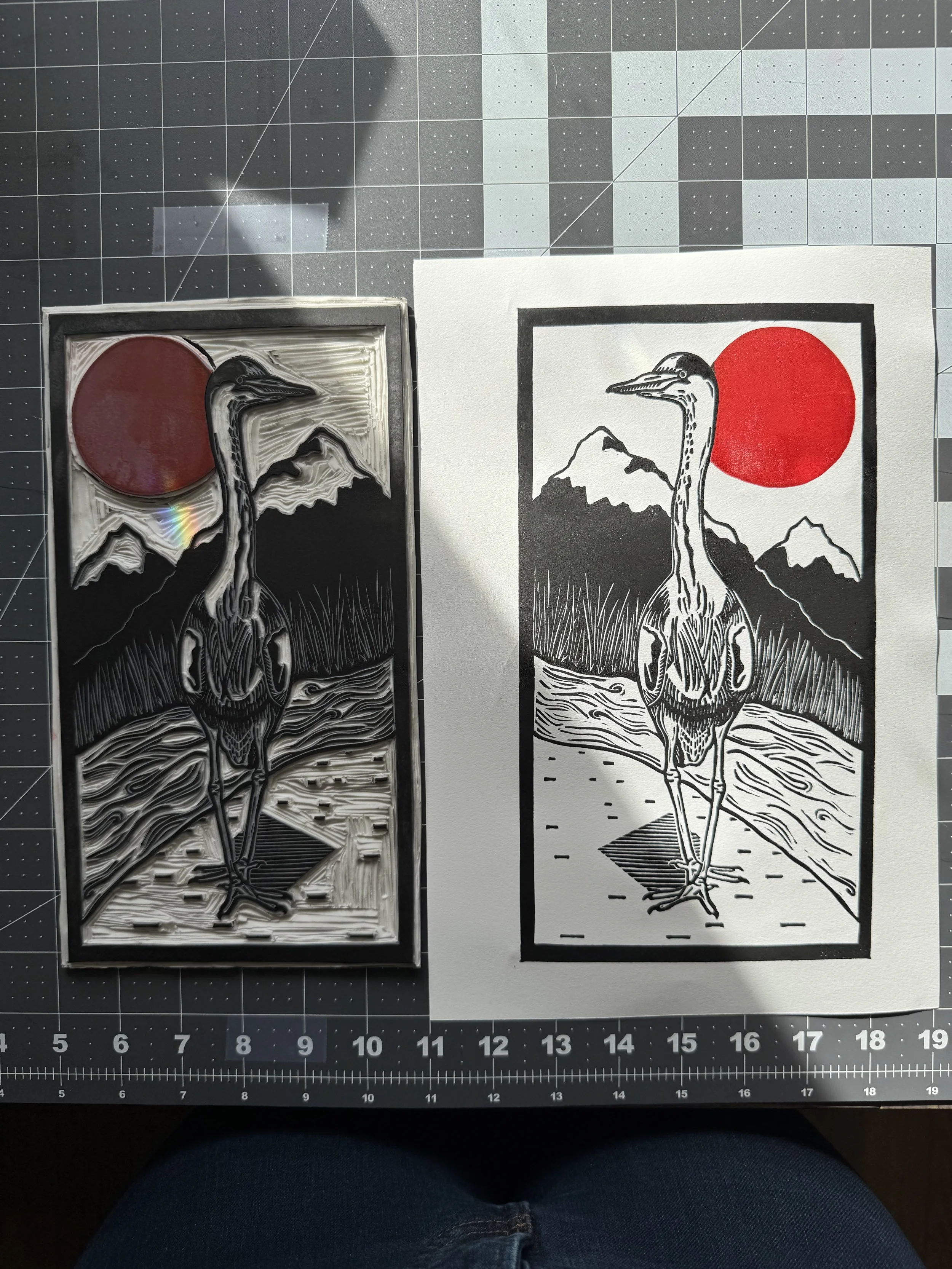 A black and red linoprint of a heron standing in front of a river, reeds, and mountains with a red sun hanging in the sky next to the carved linoleum block matching in a mirror image