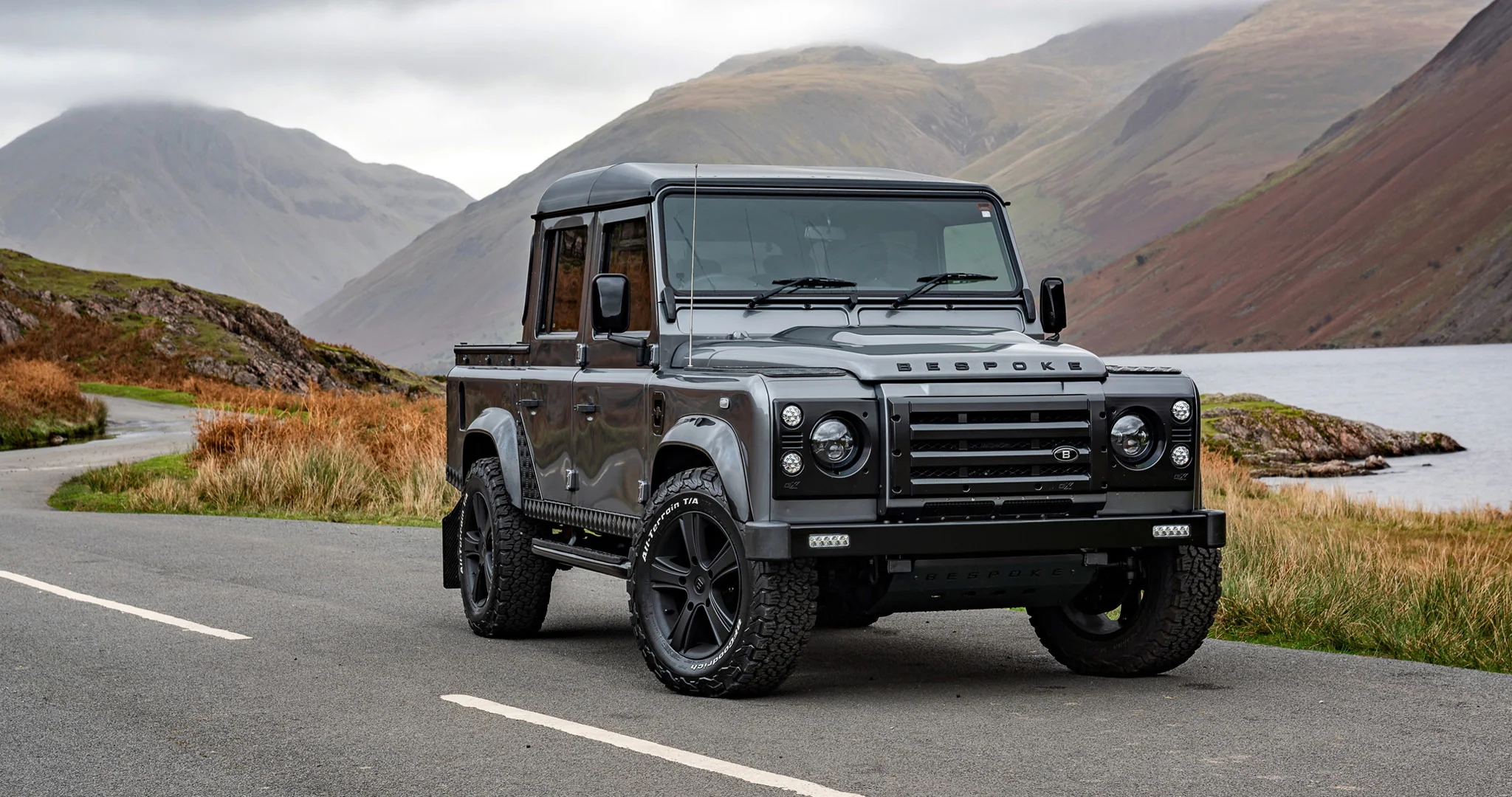 Grey customised Land Rover Defender 110 double-cab pickup photographed from the front on a mountain road beside Wast Water in the Lake District.