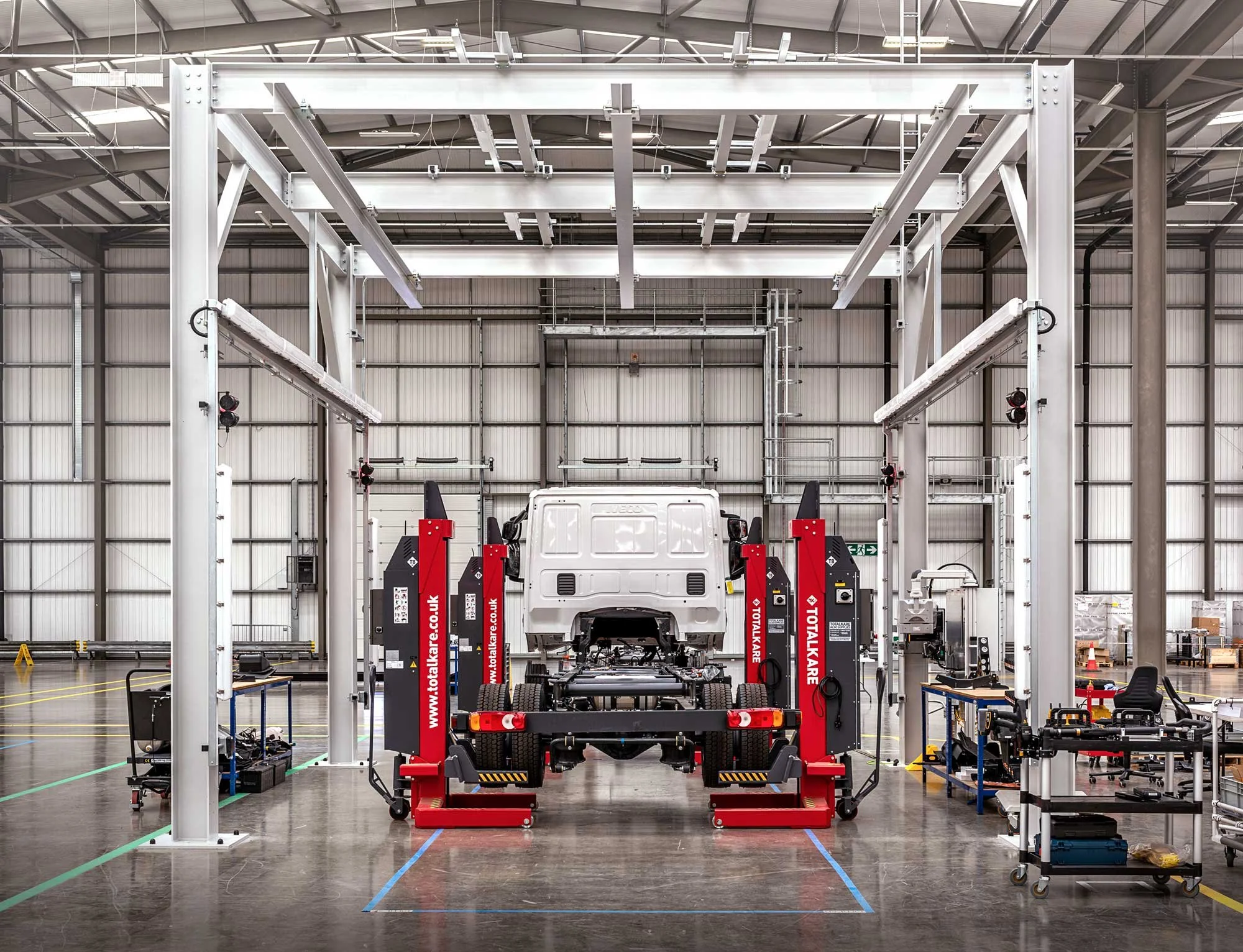 Electric truck elevated on red assembly jacks inside large hangar-style manufacturing facility – industrial manufacturing photography London