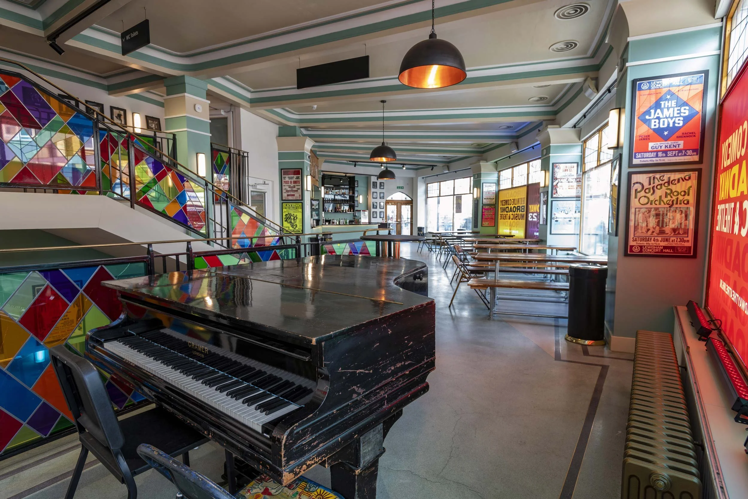 Broadway Theatre foyer and bar in Catford, featuring a piano, colourful staircase panels, wooden tables and large windows.