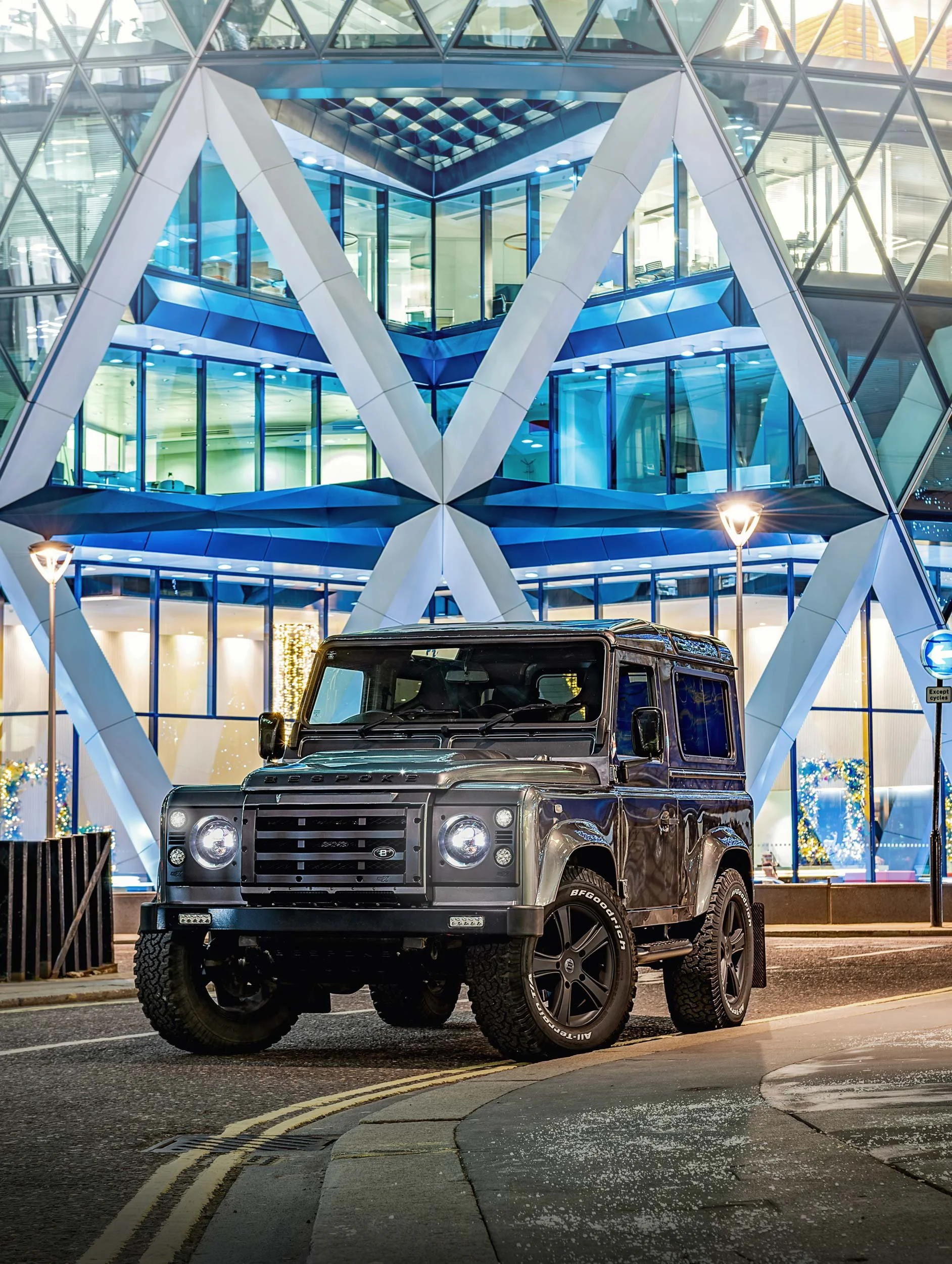 Grey bespoke Land Rover Defender 90 photographed in front of a blue-lit glass building with large geometric structural beams.