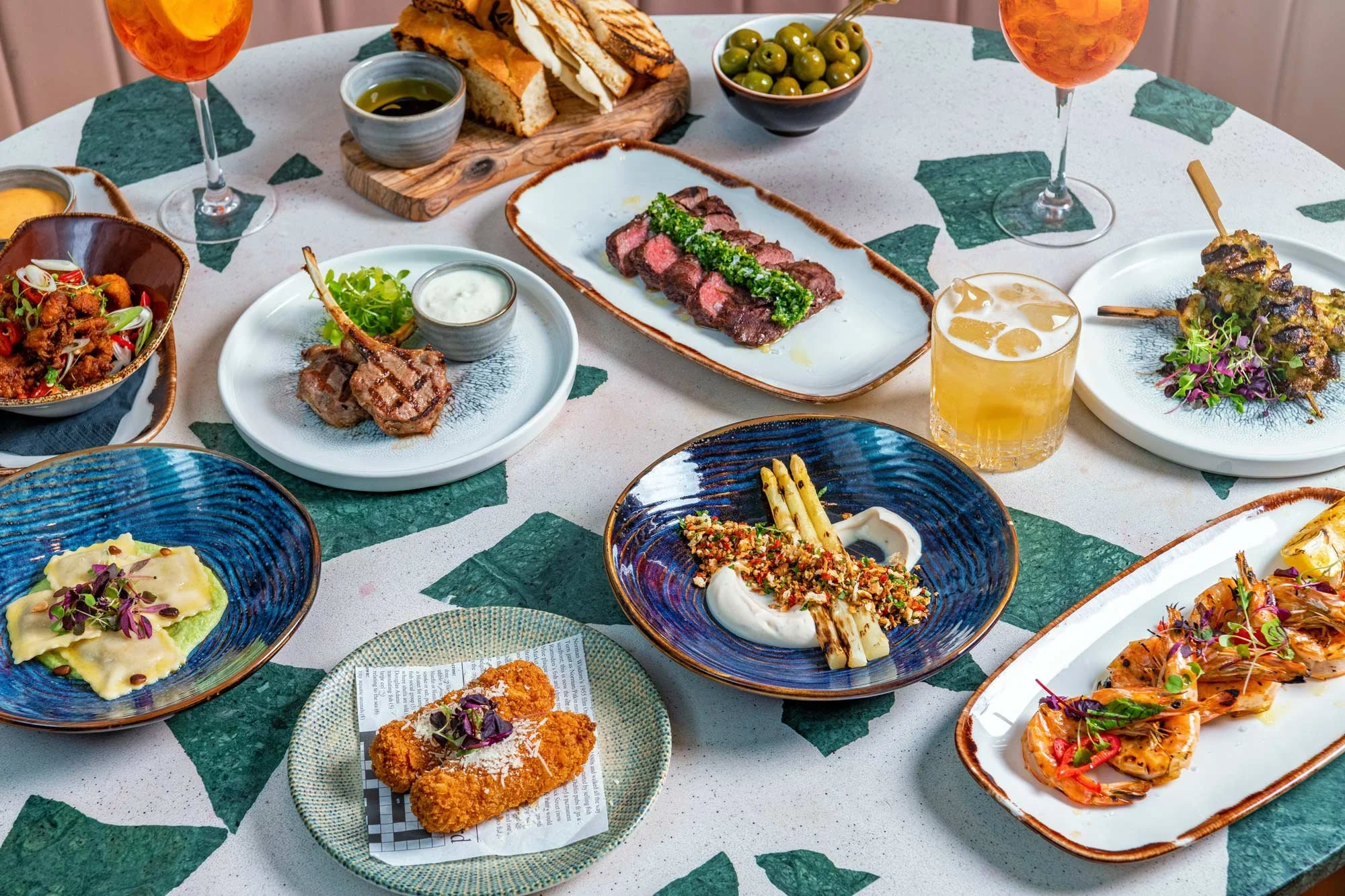 Close-up of a terrazzo table at Shakedown Restaurant in The Dixon Hotel featuring a generous spread of dishes including croquettes, fillet steak, lamb chops, ravioli and sides – London hotel restaurant food photography.