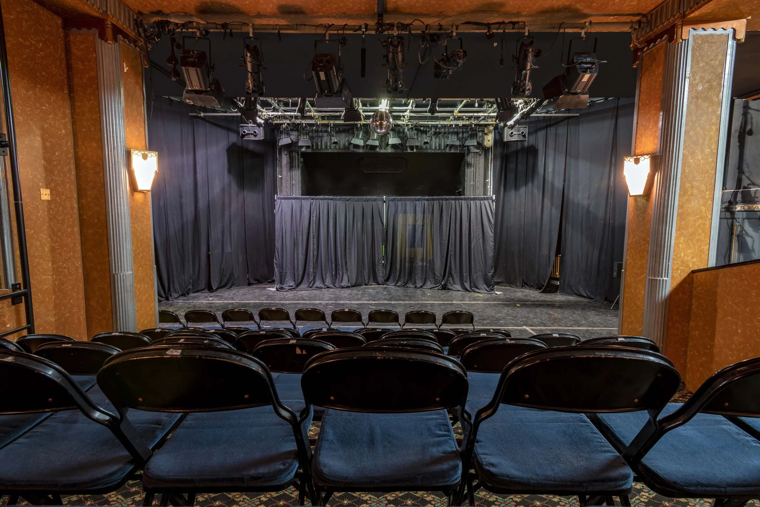 Broadway Theatre studio performance space with black stage curtains, rows of audience chairs and overhead stage lighting.