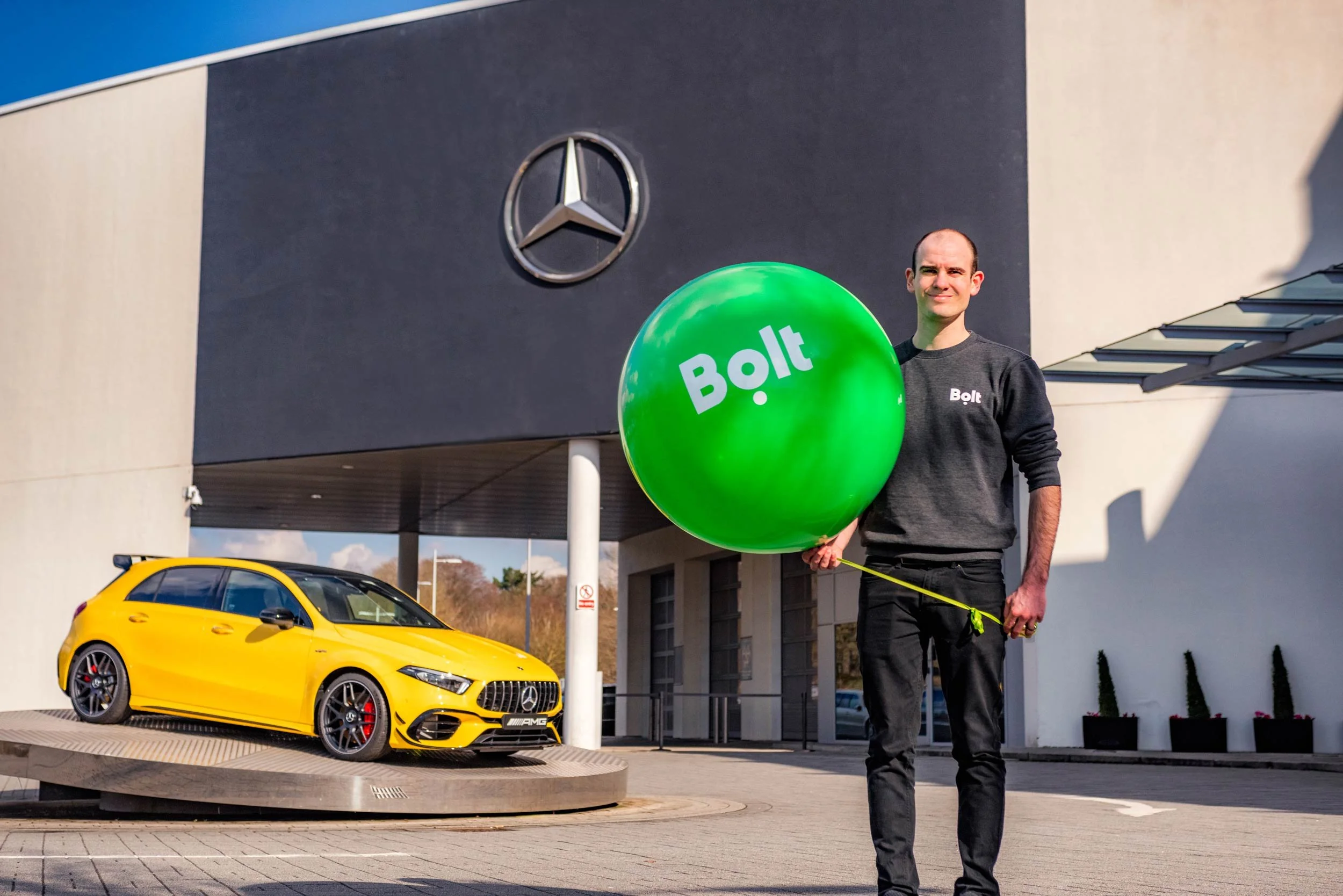 Bolt team member with a branded balloon outside Mercedes-Benz World next to a yellow Mercedes-AMG A45
