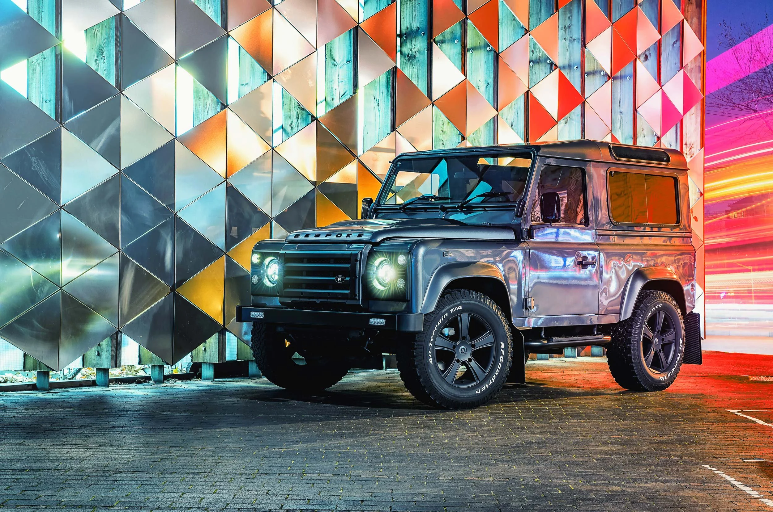 Custom grey Land Rover Defender 90 photographed at night beside a colourful geometric wall, with reflections across its bodywork.