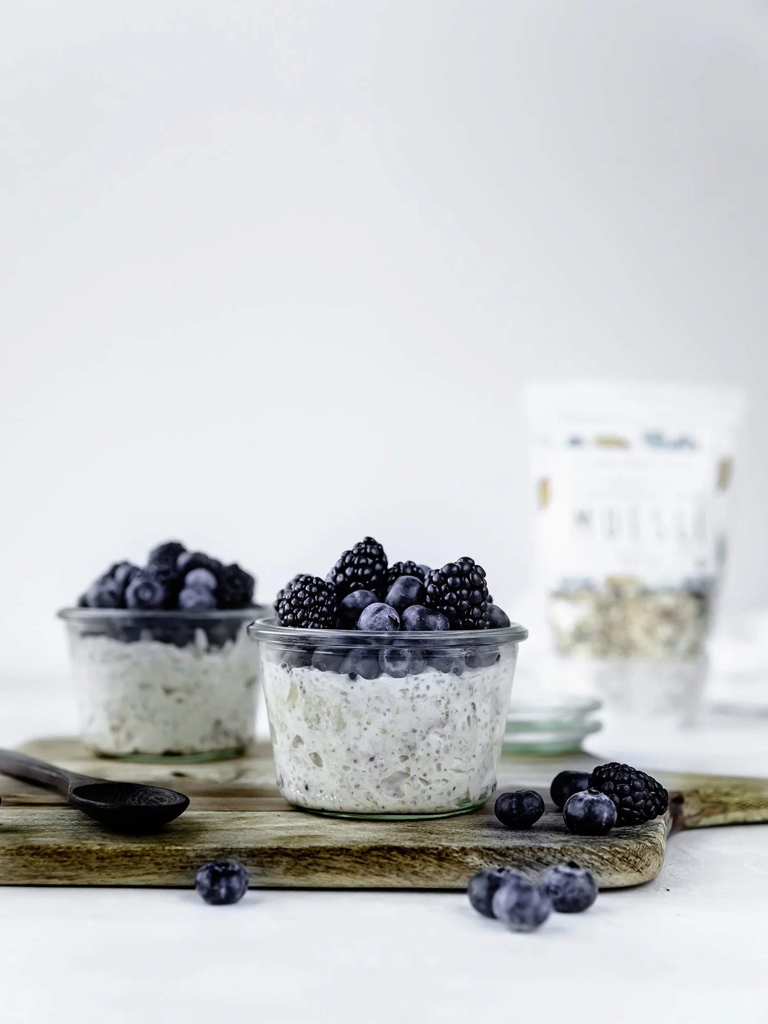 Two jars filled with overnight muesli with berries on top next to a bag of Victory Dance Foods Blueberry Muesli.