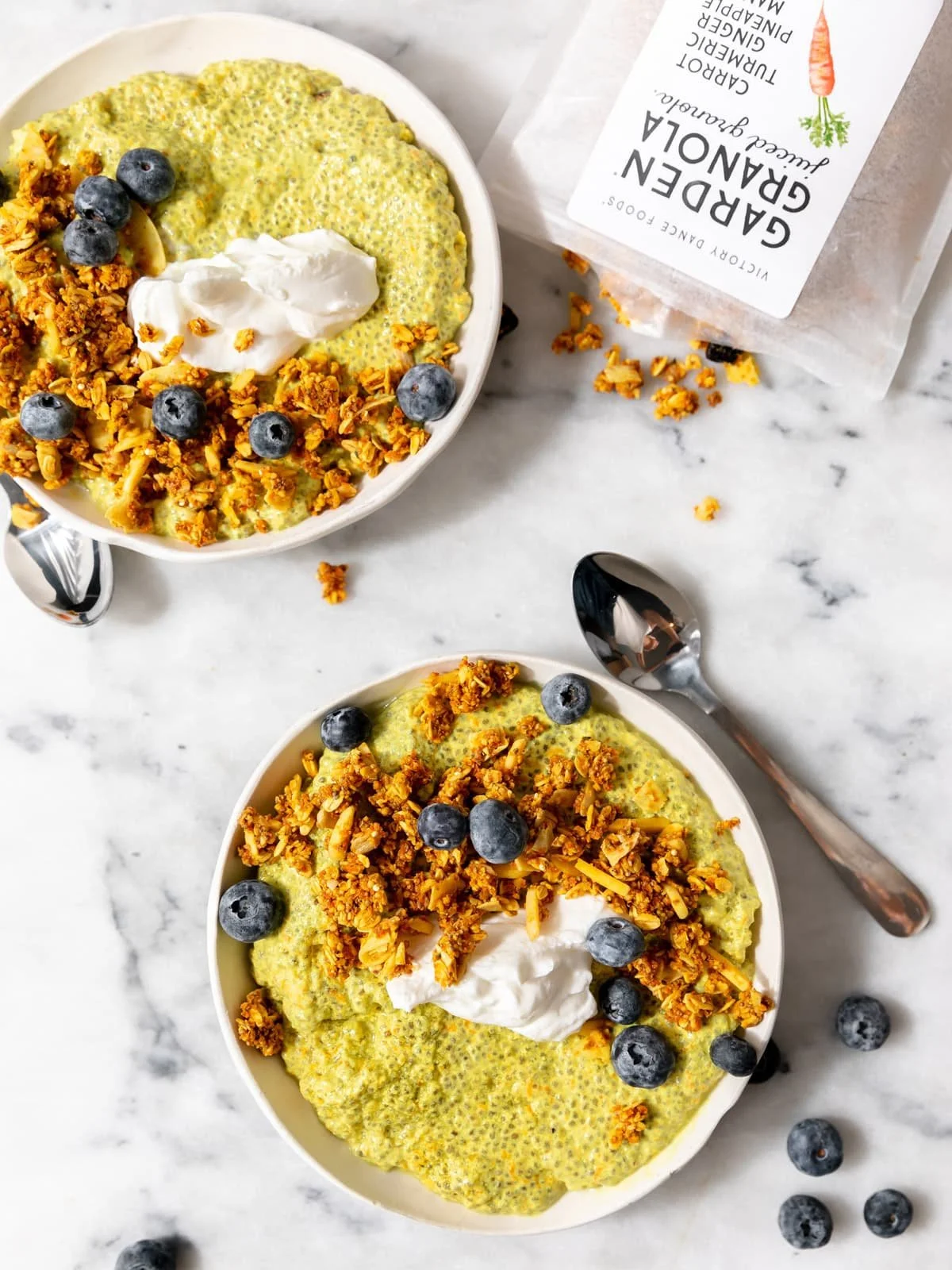 Overhead view of two golden turmeric chia pudding bowls topped with Carrot Turmeric Garden Granola, coconut yogurt, and fresh blueberries on white marble surface
