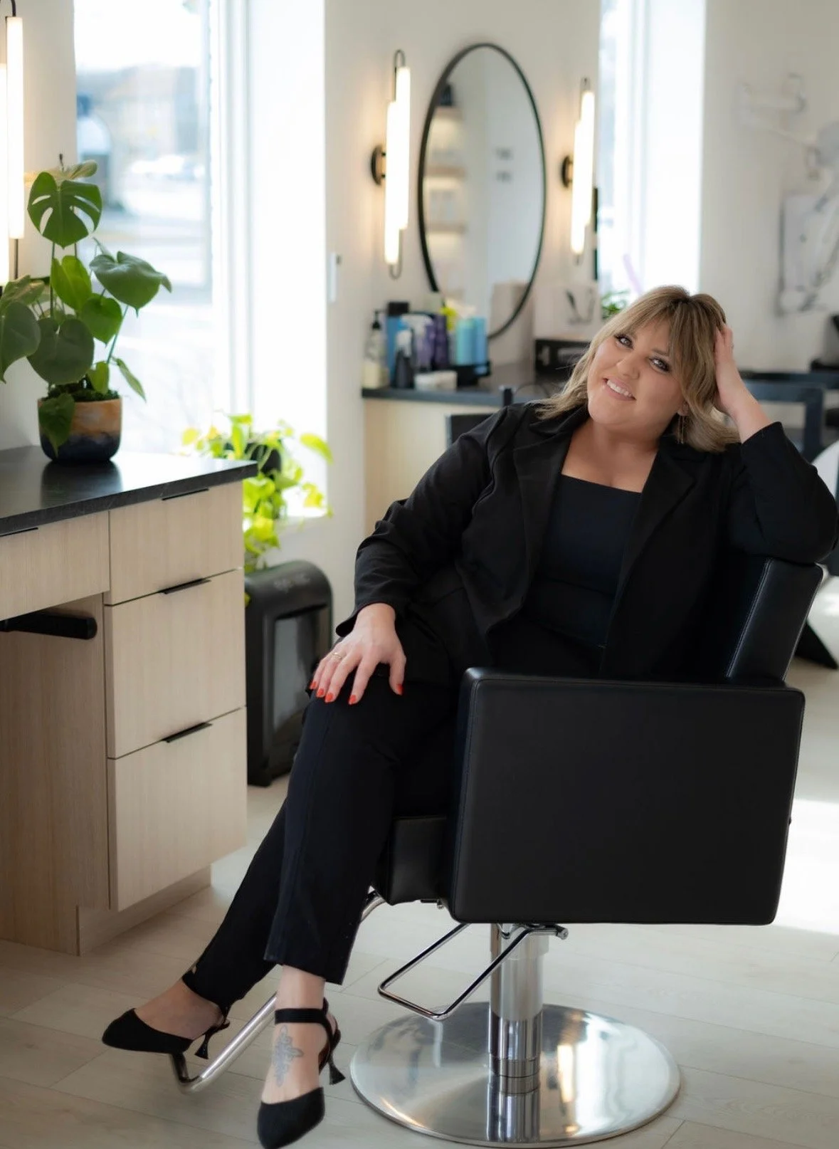 Carmen Grapentine sitting in a hair-styling chair wearing all black and with a hand in her hair, smiling.