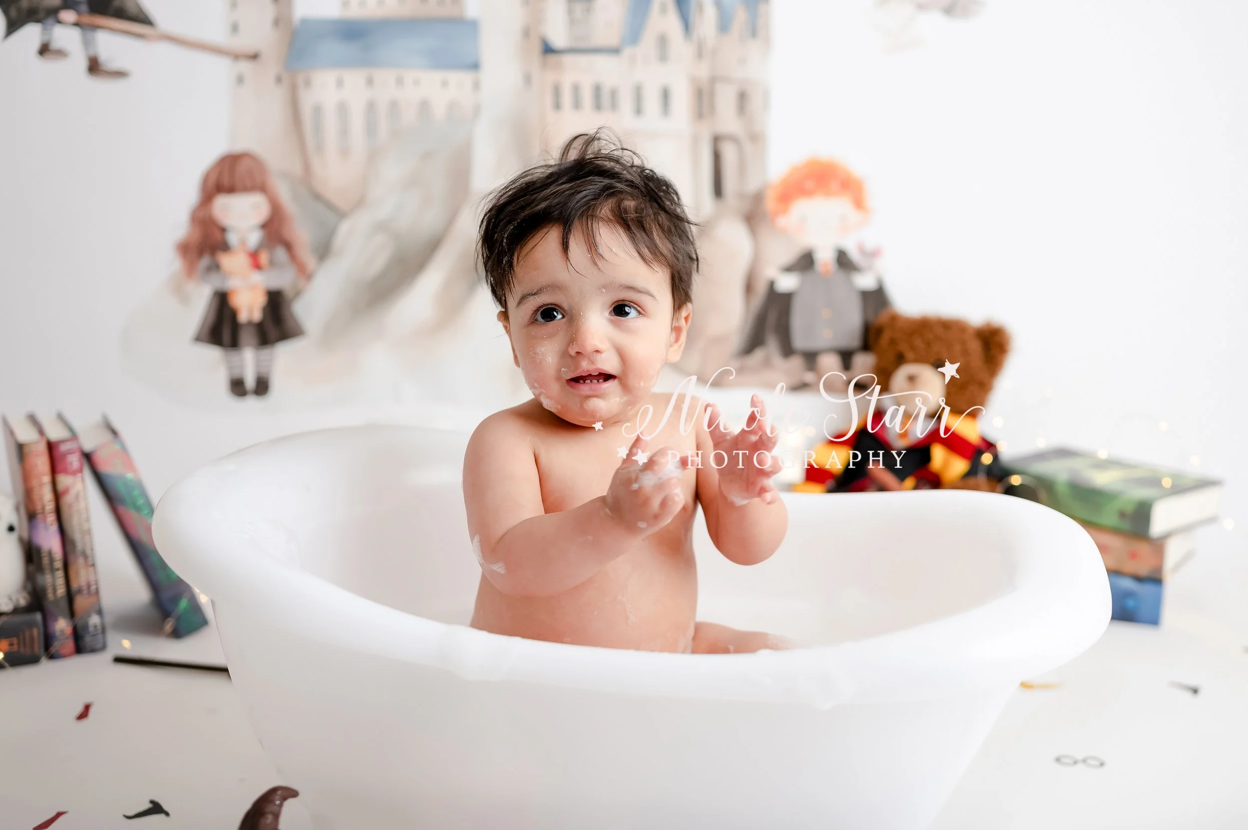baby claps sitting in white tub in front of watercolor Hogwarts backdrop during light & airy Harry Potter theme cake smash photographed by Saratoga Springs baby photographer Nicole Starr Photography