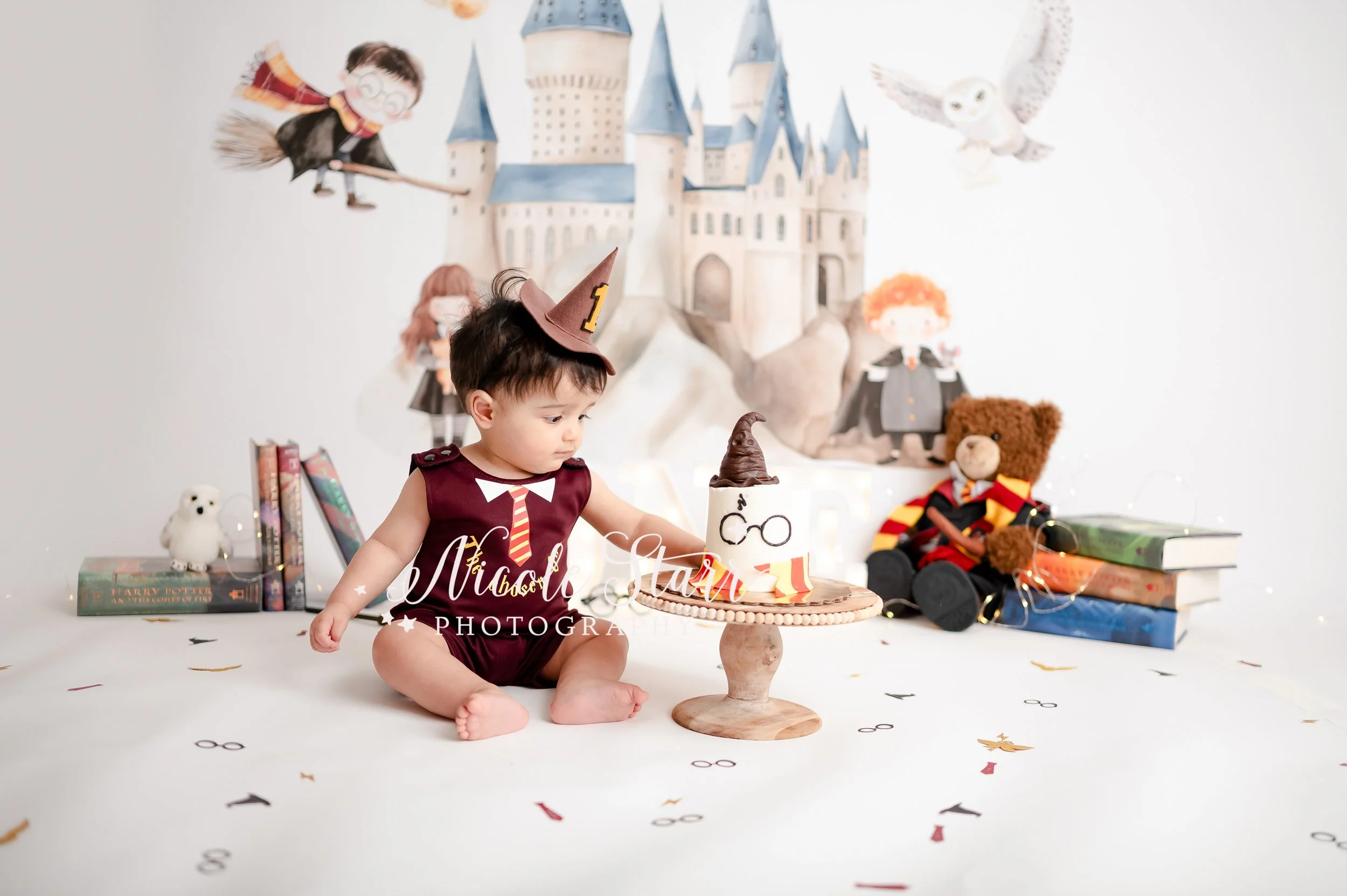 baby in red and yellow onesie reaches for cake in front of watercolor Hogwarts backdrop during light & airy Harry Potter theme cake smash photographed by Saratoga Springs baby photographer Nicole Starr Photography