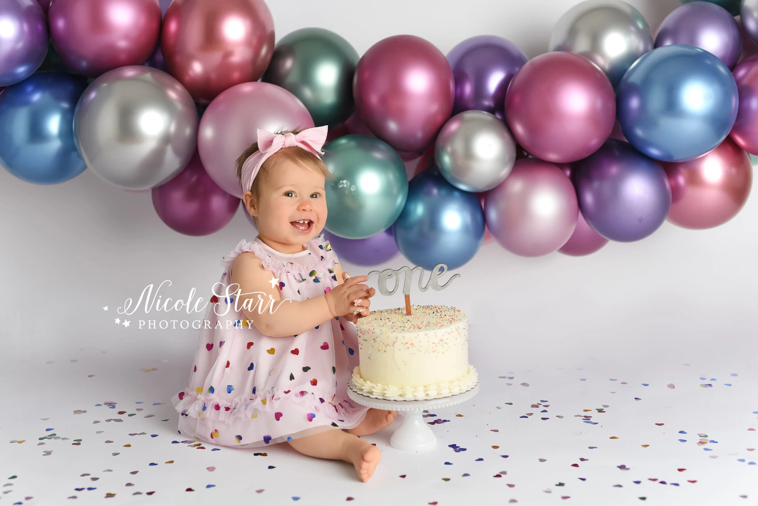 baby girl plays with cake during cake smash with balloon garland hanging behind her photographed by Delmar NY cake smash photographer Nicole Starr Photography