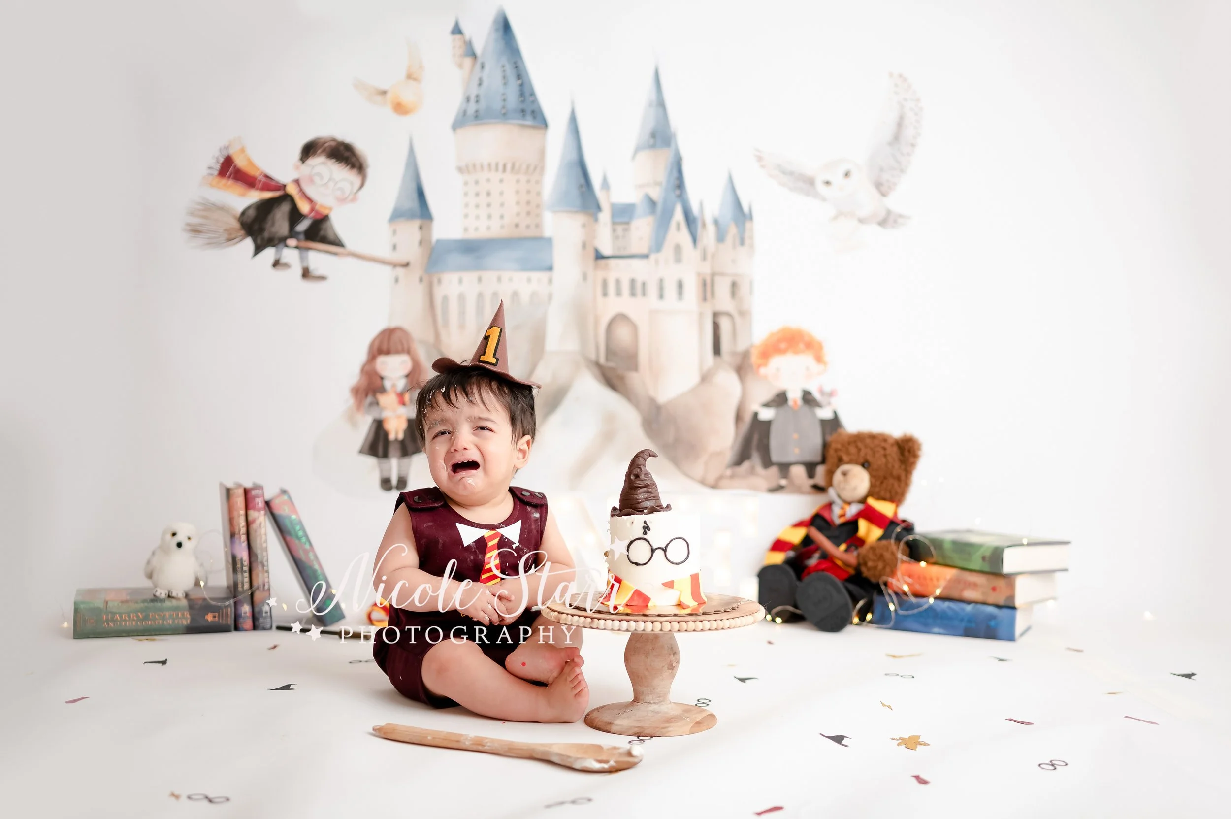 baby fusses sitting in onesie that says “the chosen one” in front of watercolor Hogwarts backdrop during light & airy Harry Potter theme cake smash photographed by Saratoga Springs baby photographer Nicole Starr Photography