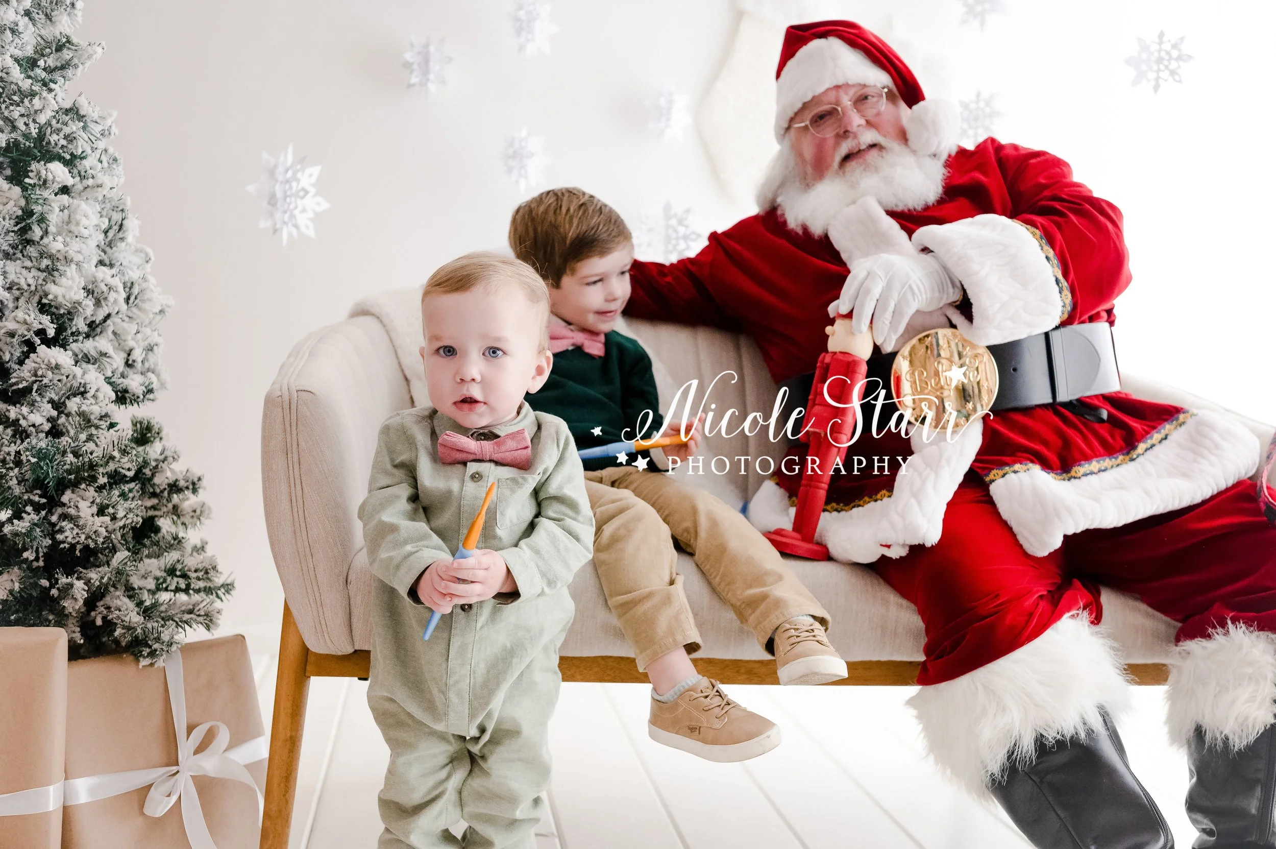 Santa paints nutcracker with brothers in green sweaters on sofa during Saratoga Springs holiday studio session photographed by NY children’s photographer Nicole Starr Photography