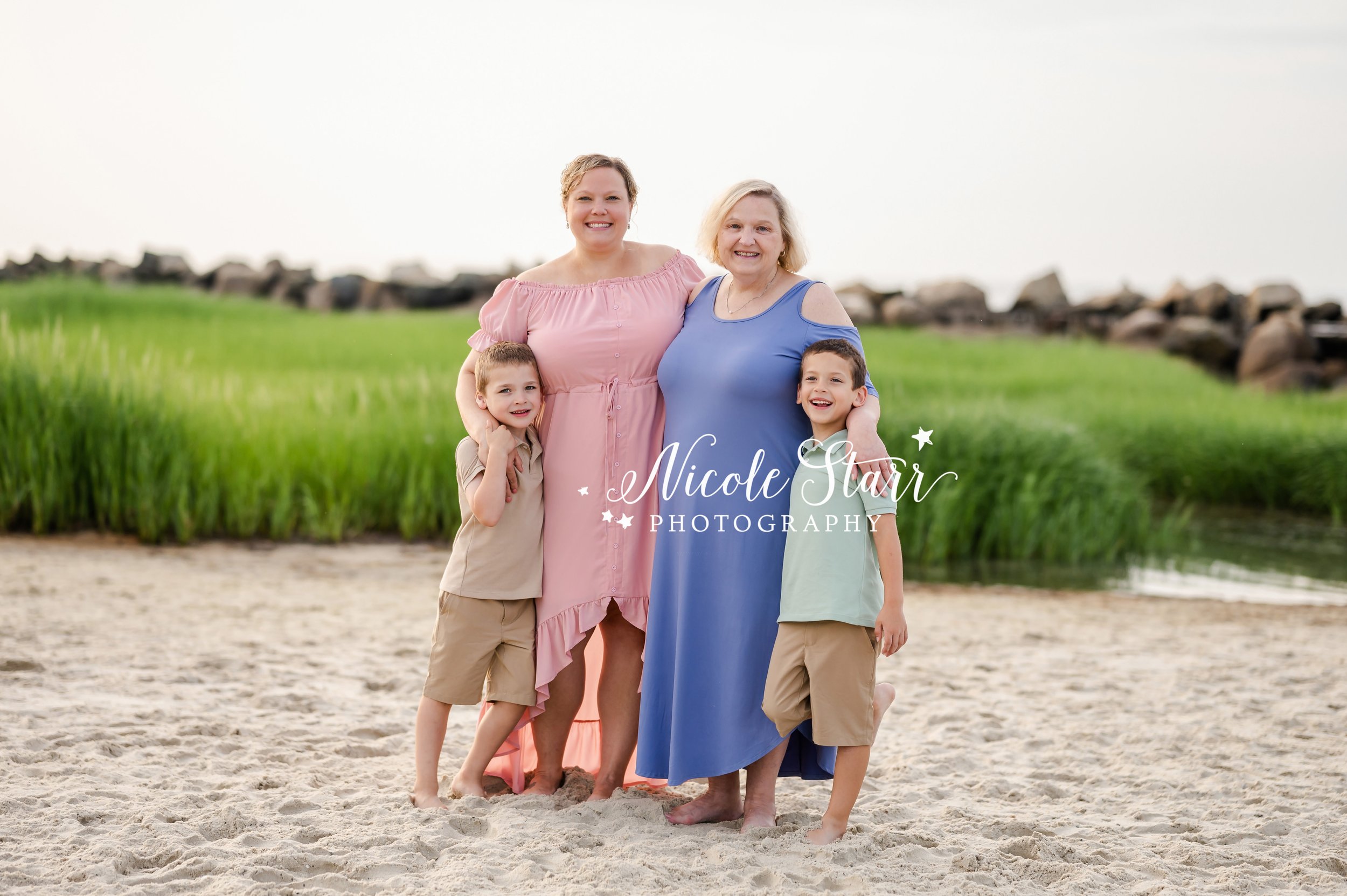 woman in pink dress hugs mom in purple dress with two sons in front of sea grass during summer Cape Cod extend family portraits with MA beach photographer Nicole Starr Photography
