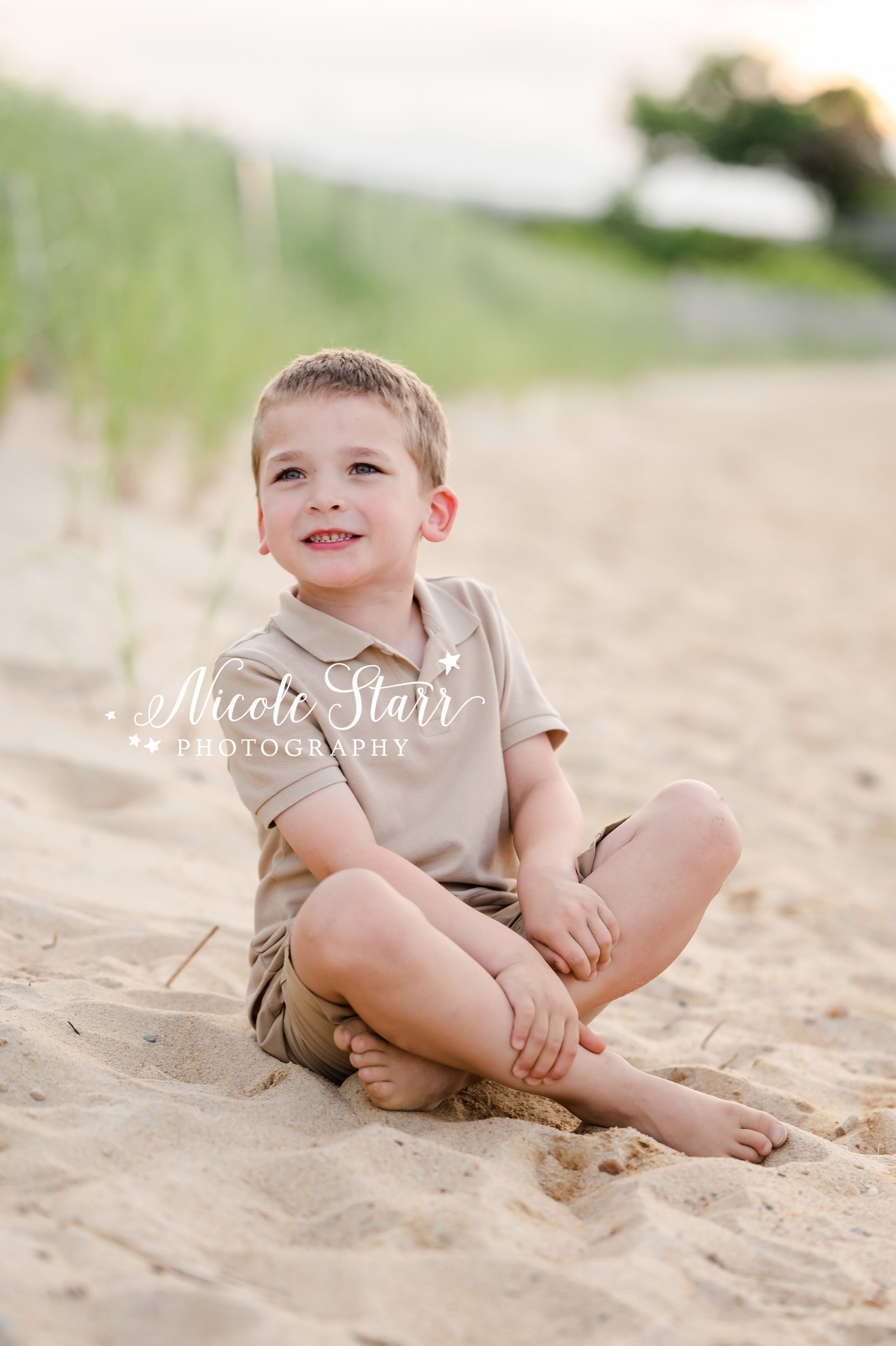 boy in tan polo sits on dune by sea grass on beach during summer Cape Cod extend family portraits with MA beach photographer Nicole Starr Photography