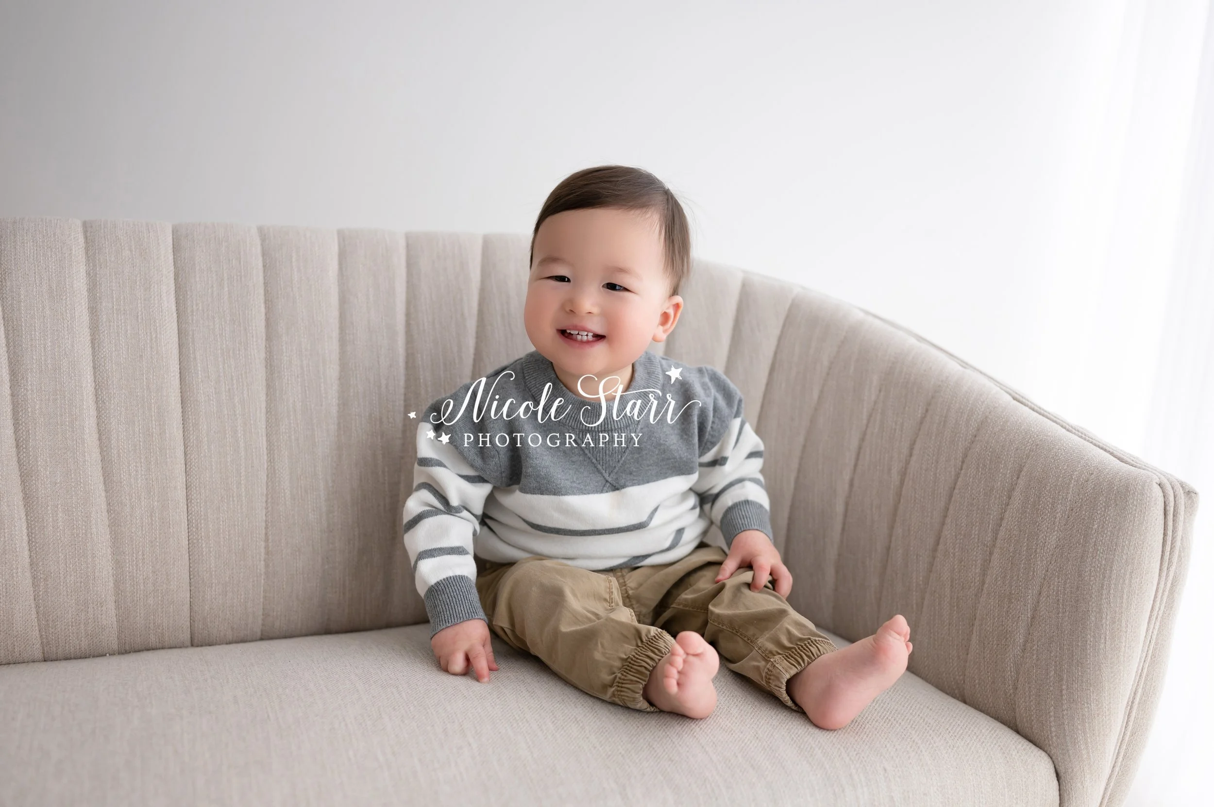 toddler sits on tan sofa with white and grey sweater during photos with  Saratoga Springs NY family photographer Nicole Starr Photography