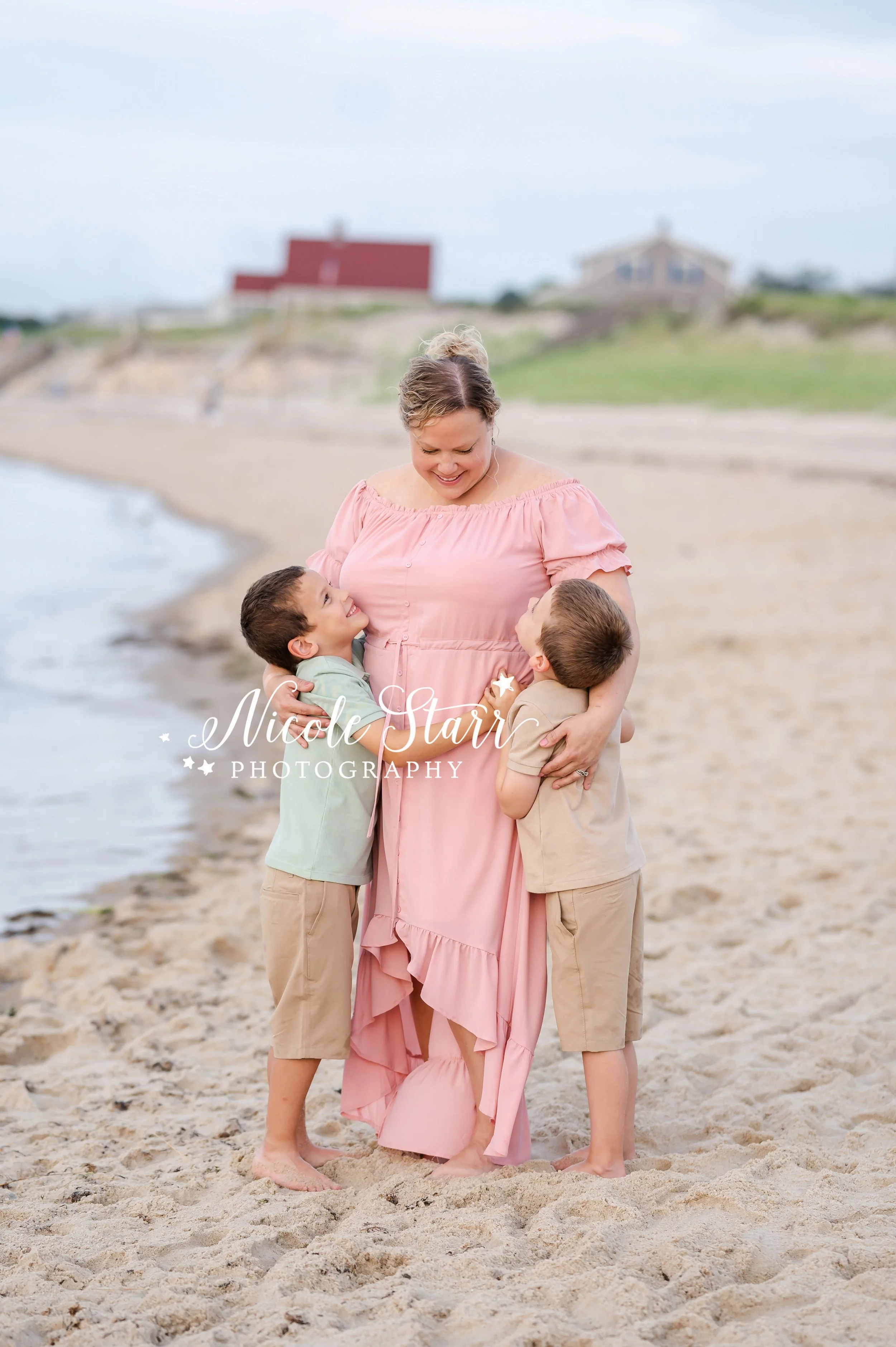 boys hug mom in pink gown standing on sand on beach along water during summer family portraits with MA beach photographer Nicole Starr Photography