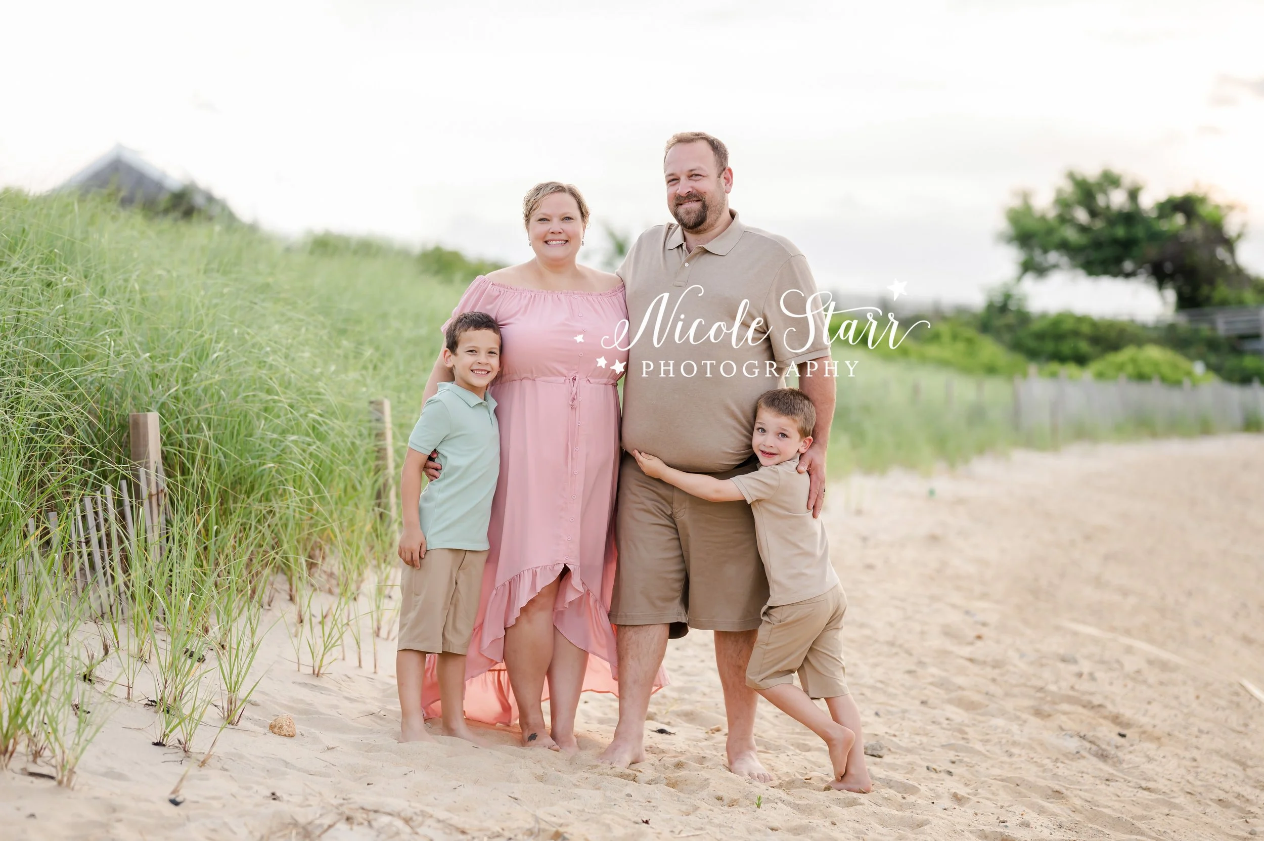 boys hug parents standing by sea grass and wooden fence on beach during summer Cape Cod extend family portraits with MA beach photographer Nicole Starr Photography