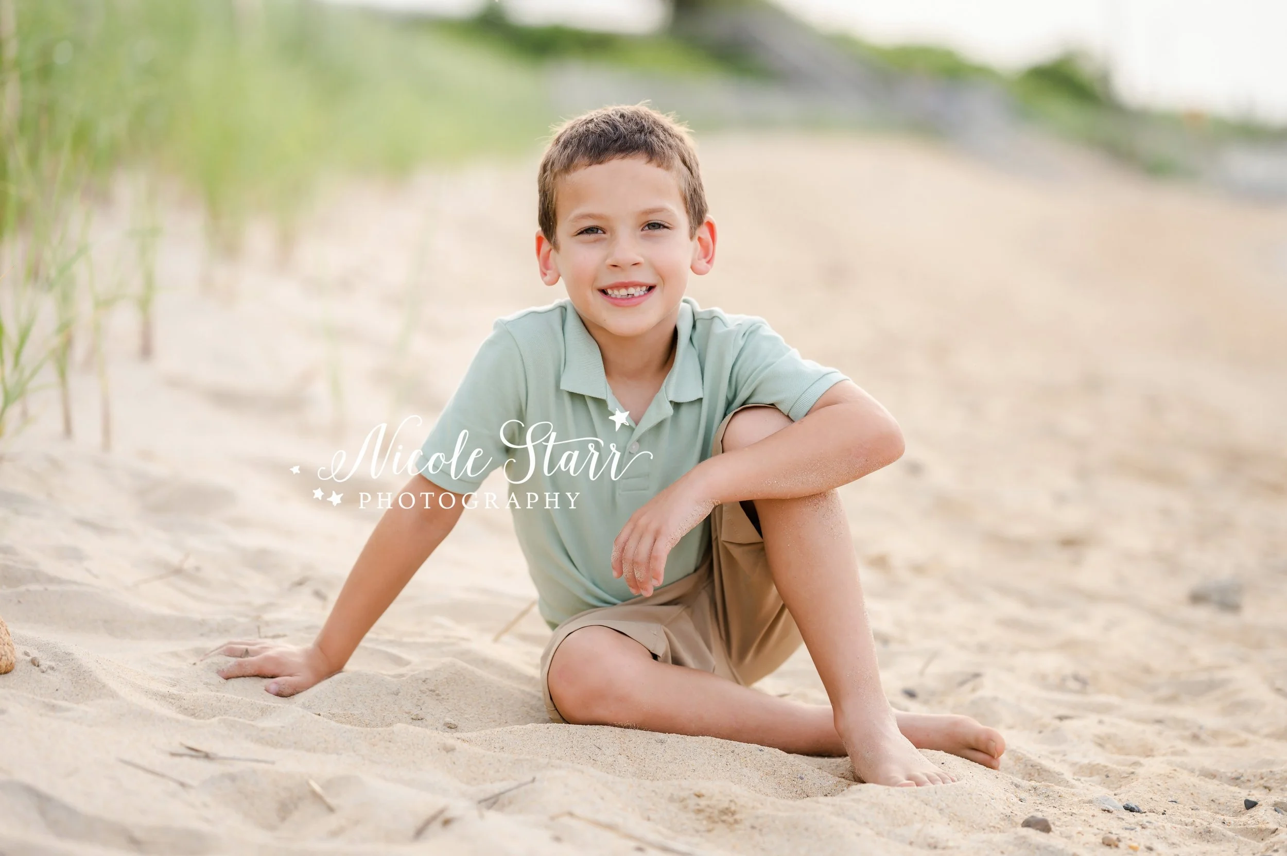 boy in sea green polo sits on dune by sea grass on beach during summer Cape Cod extend family portraits with MA beach photographer Nicole Starr Photography