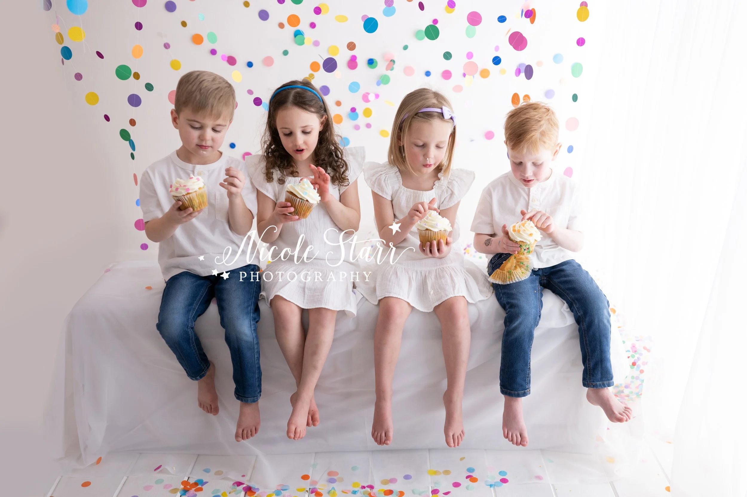 four toddlers in white dresses and white shirts with jeans sit eating cupcakes in studio with rainbow streamers for cake smash with Nicole Starr Photography, Saratoga Springs children’s photographer