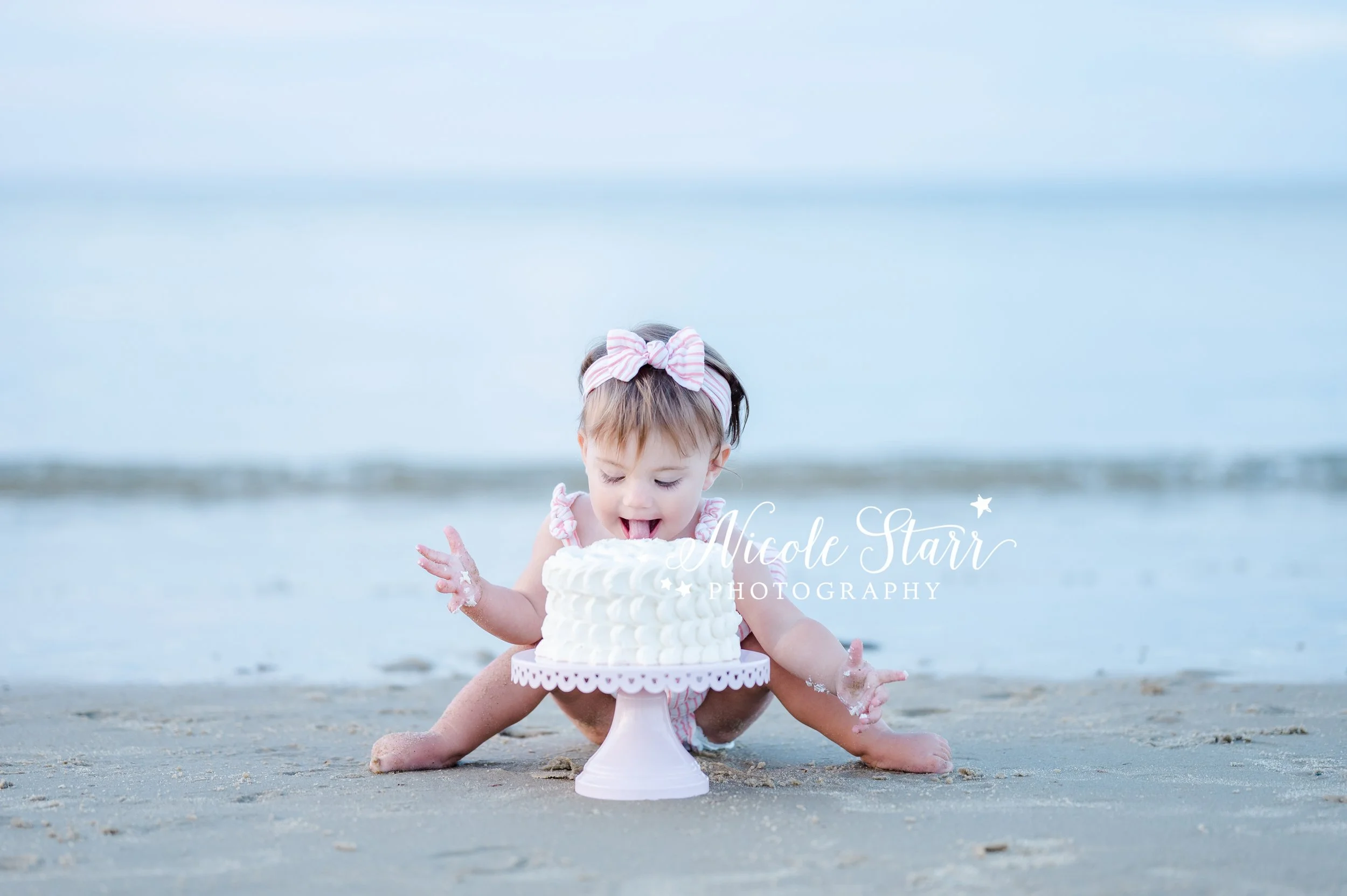 baby girl licks white cake on pink cake stand in front of the ocean during cake smash on Cape Cod with Cape Cod MA cake smash photographer Nicole Starr Photography