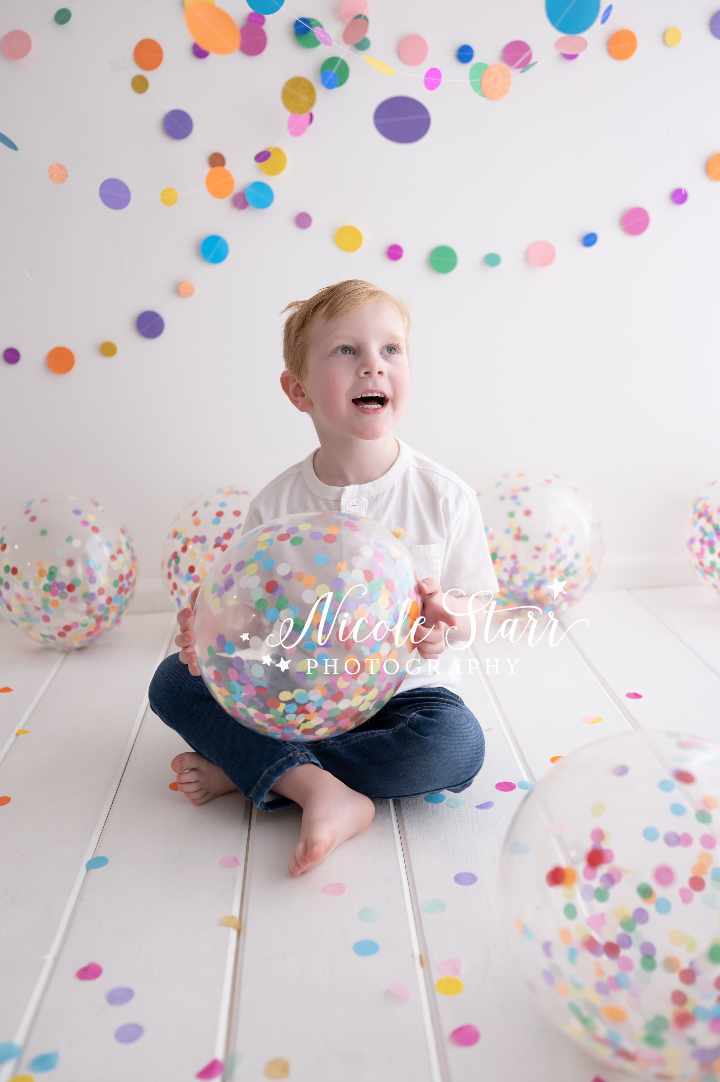 fine year old boy in white shirt and jeans sits on studio floor holding confetti beach balls in front of rainbow streamers for cake smash with Nicole Starr Photography, Saratoga Springs children’s photographer