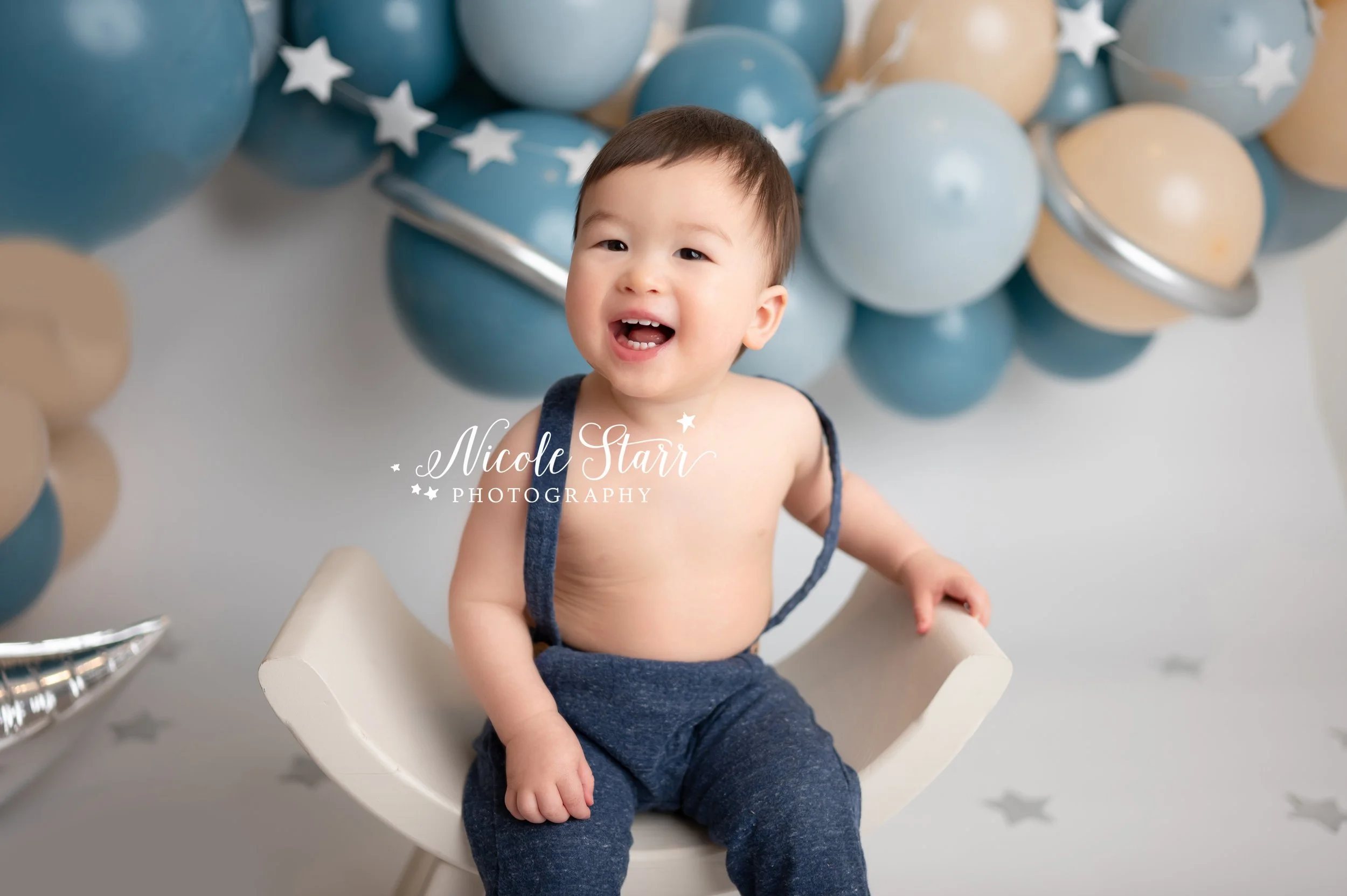 toddler in blue suspenders sits in chair in front of blue space themed backdrop during cake smash with Nicole Starr Photography