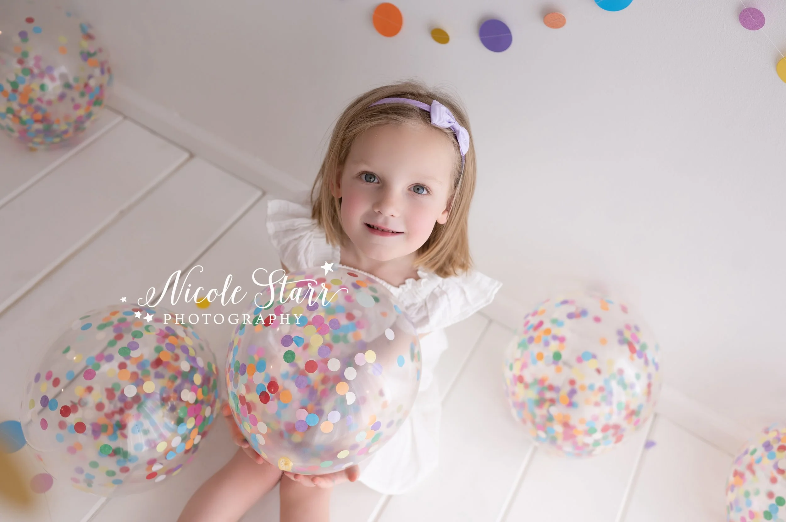 blonde girl in white holds beach ball in studio with confetti beach balls in front of rainbow streamers for cake smash with Nicole Starr Photography, Saratoga Springs children’s photographer