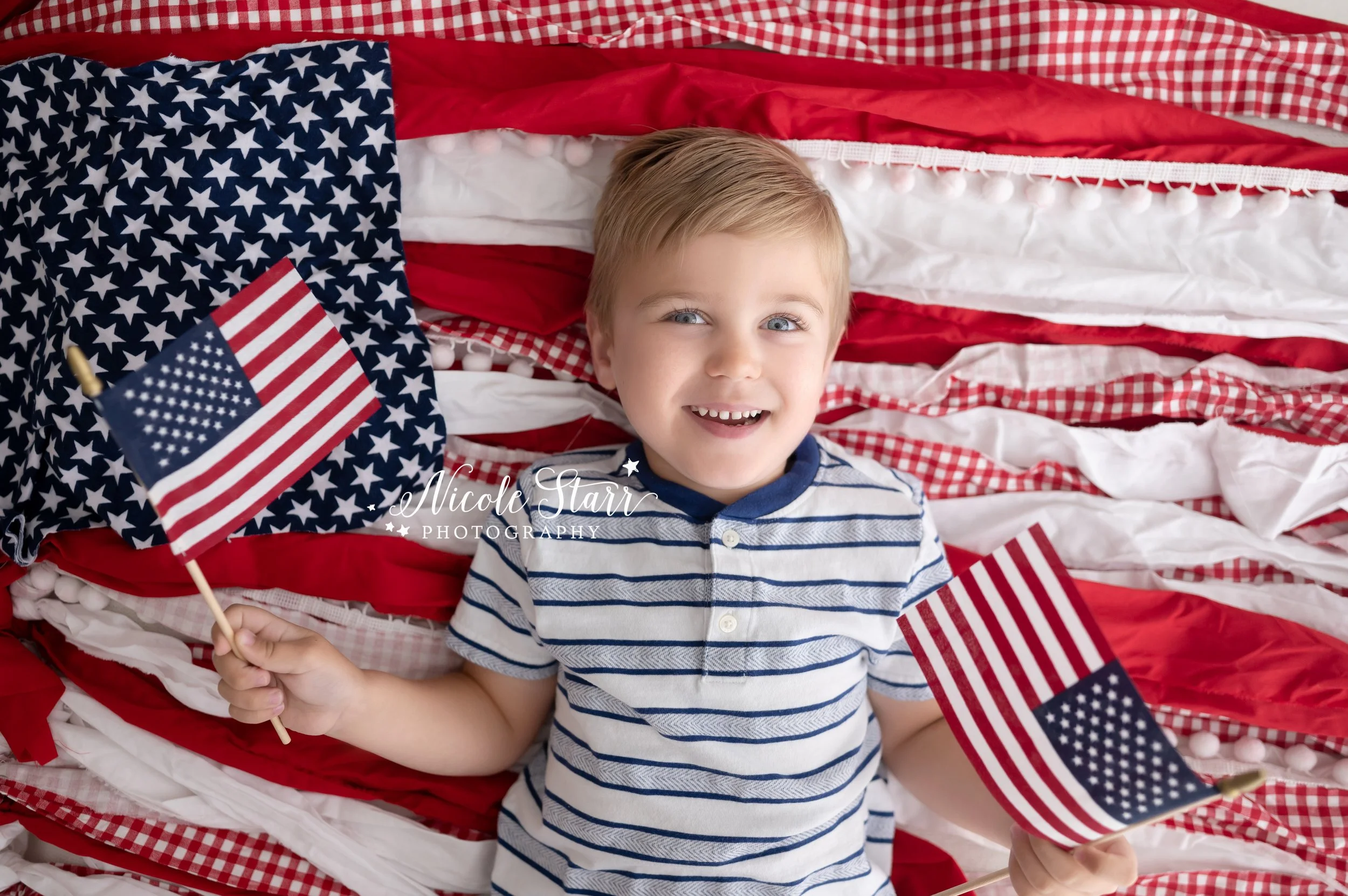 toddler holds American flags laying on red and white backdrop during 4th of July portraits in Saratoga Springs studio
