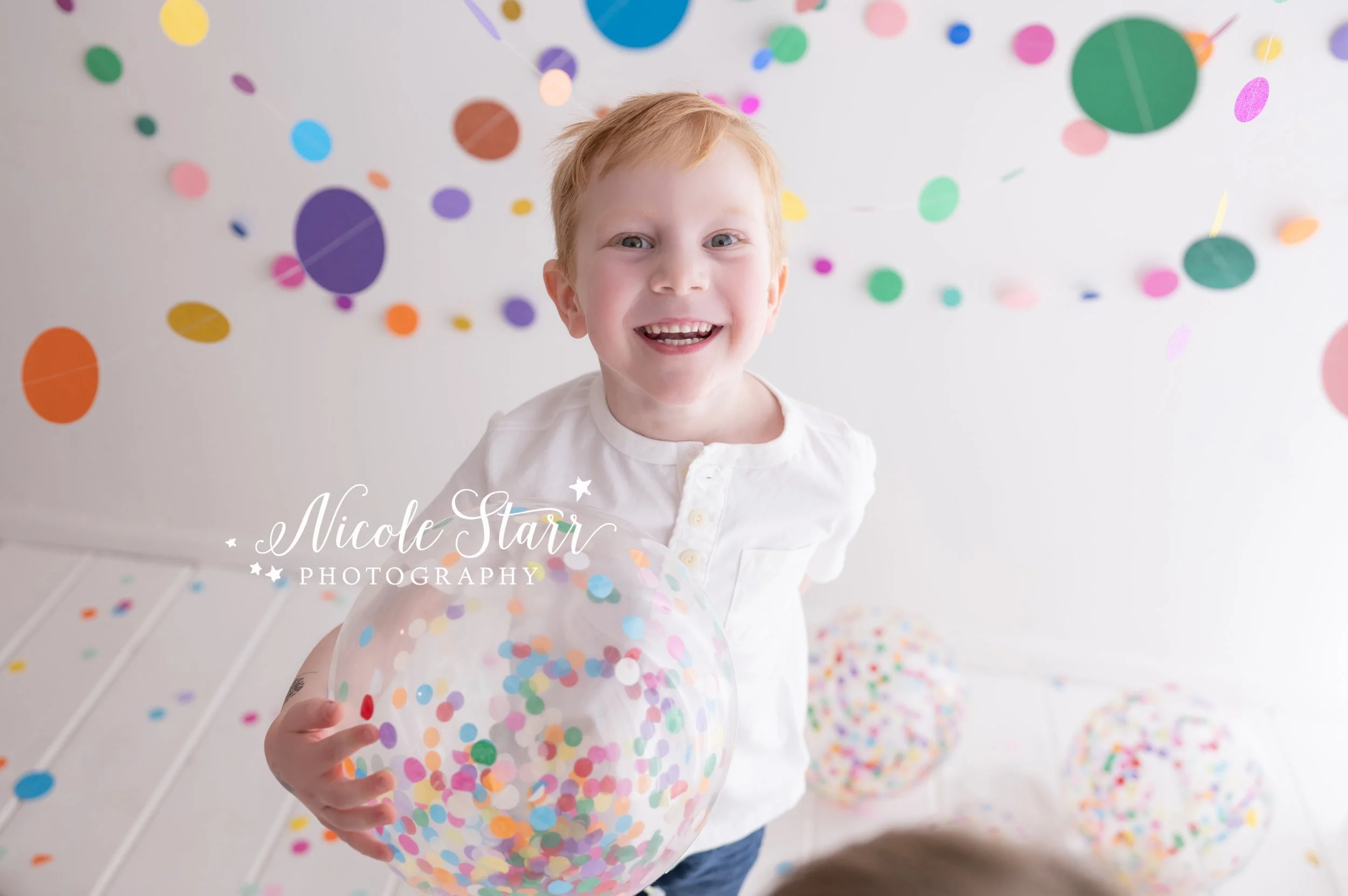 boy in white shirt laughs holding confetti beach ball in studio with rainbow streamers for cake smash with Nicole Starr Photography, Saratoga Springs children’s photographer