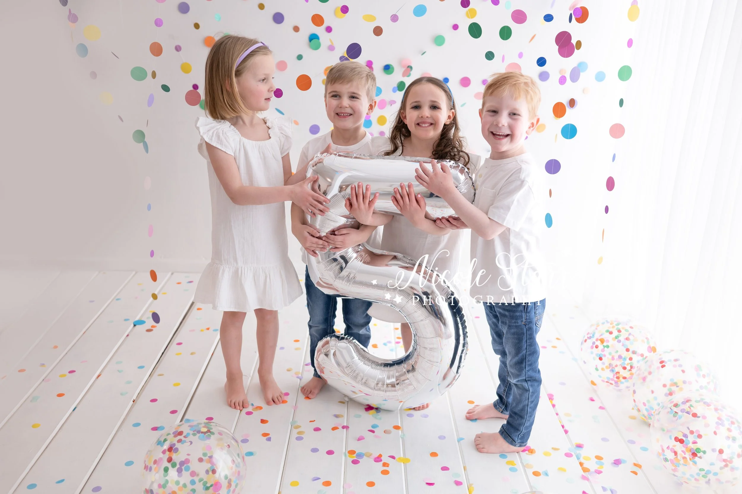 four toddlers in white dresses and white shirts with jeans pose with giant silver 5 balloon in studio with rainbow streamers for cake smash with Nicole Starr Photography, Saratoga Springs children’s photographer