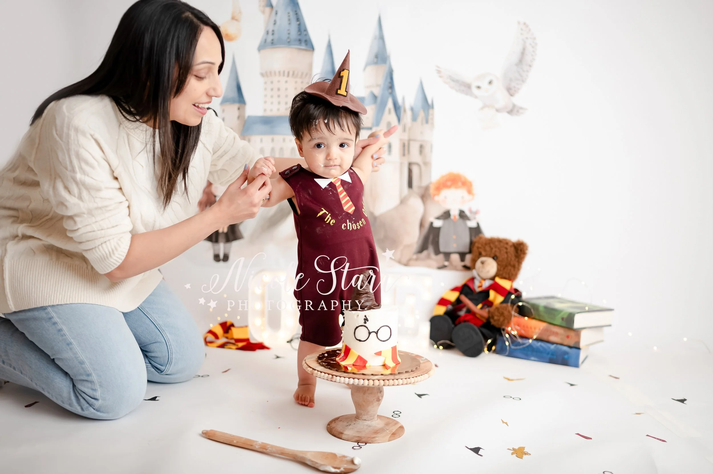 mom kneels next to son helping him stand in onesie that says “the chosen one” in front of watercolor Hogwarts backdrop during light & airy Harry Potter theme cake smash photographed by Saratoga Springs baby photographer Nicole Starr Photography