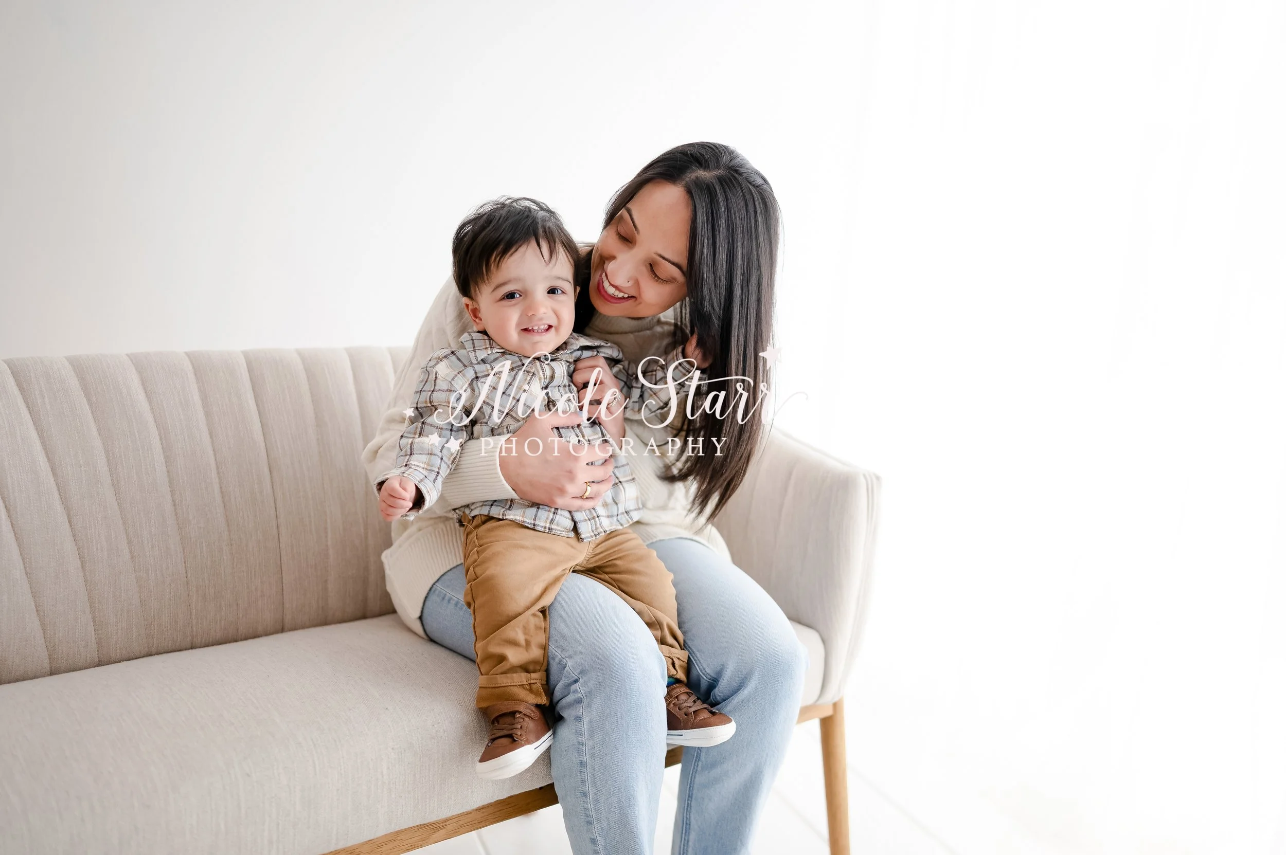mom in white sweater and jeans holds one year old in plaid shirt and brown pants on her lap in studio during first year birthday portraits with Saratoga Springs baby photographer Nicole Starr Photography
