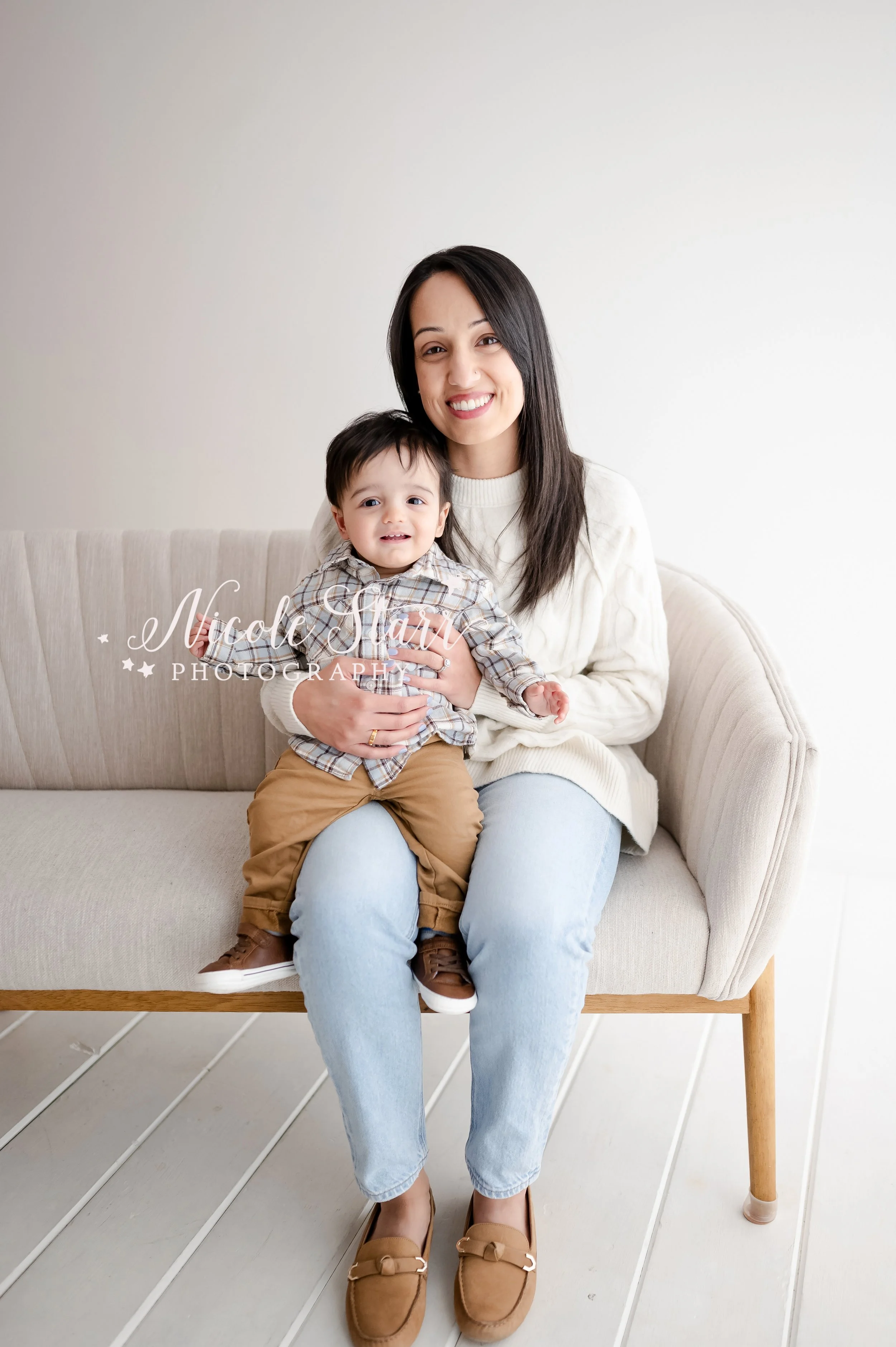 mom in white sweater and jeans holds one year old in plaid shirt and brown pants on her lap in studio during first year birthday portraits with Saratoga Springs baby photographer Nicole Starr Photography