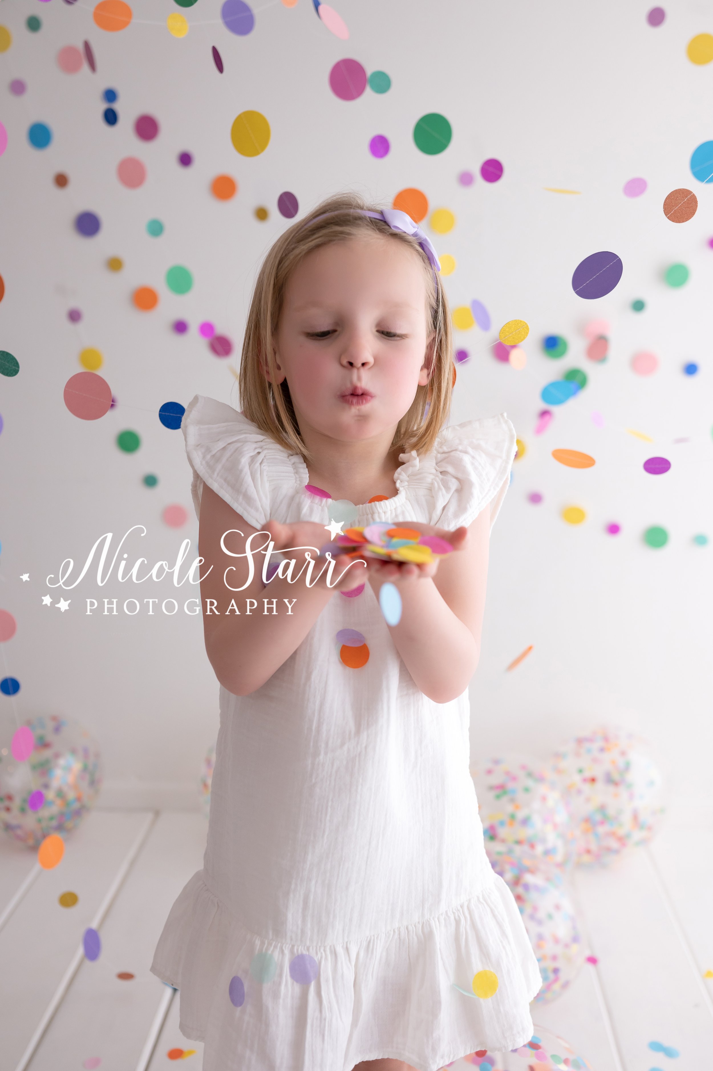 blonde girl in white dress blows confetti from hands in studio with confetti beach balls in front of rainbow streamers for cake smash with Nicole Starr Photography, Saratoga Springs children’s photographer