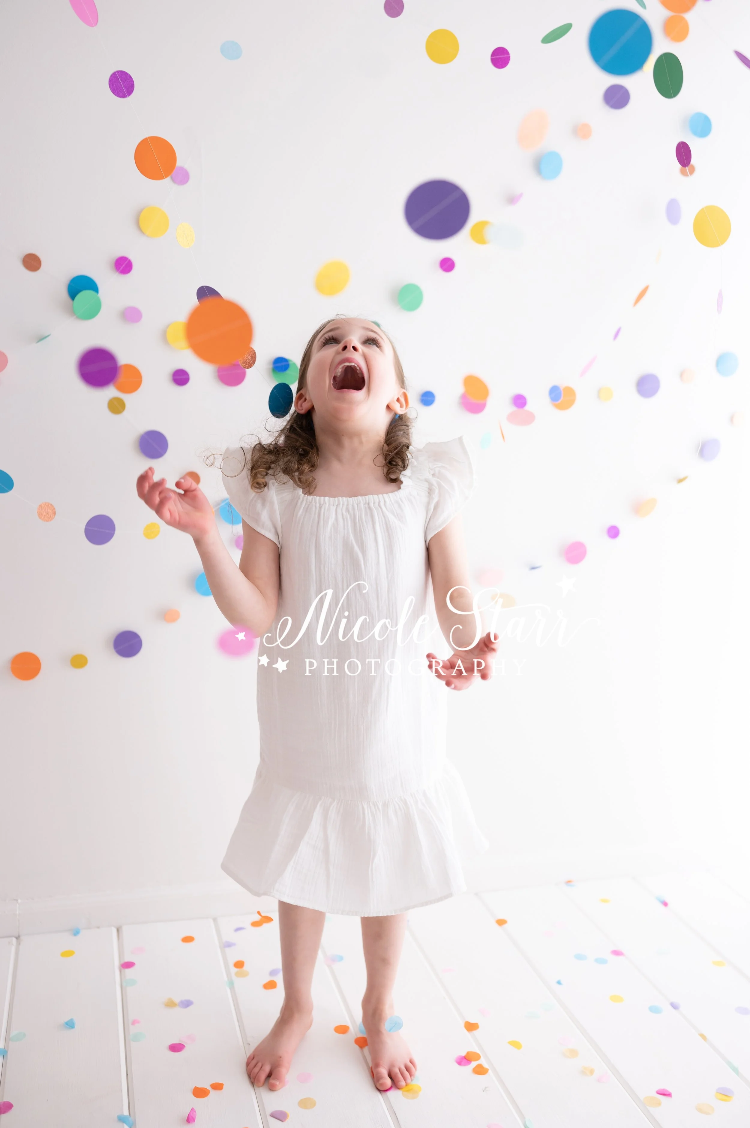 girl with brown hair in white dress throws confetti and beach balls in studio with rainbow streamers for cake smash with Nicole Starr Photography, Saratoga Springs children’s photographer
