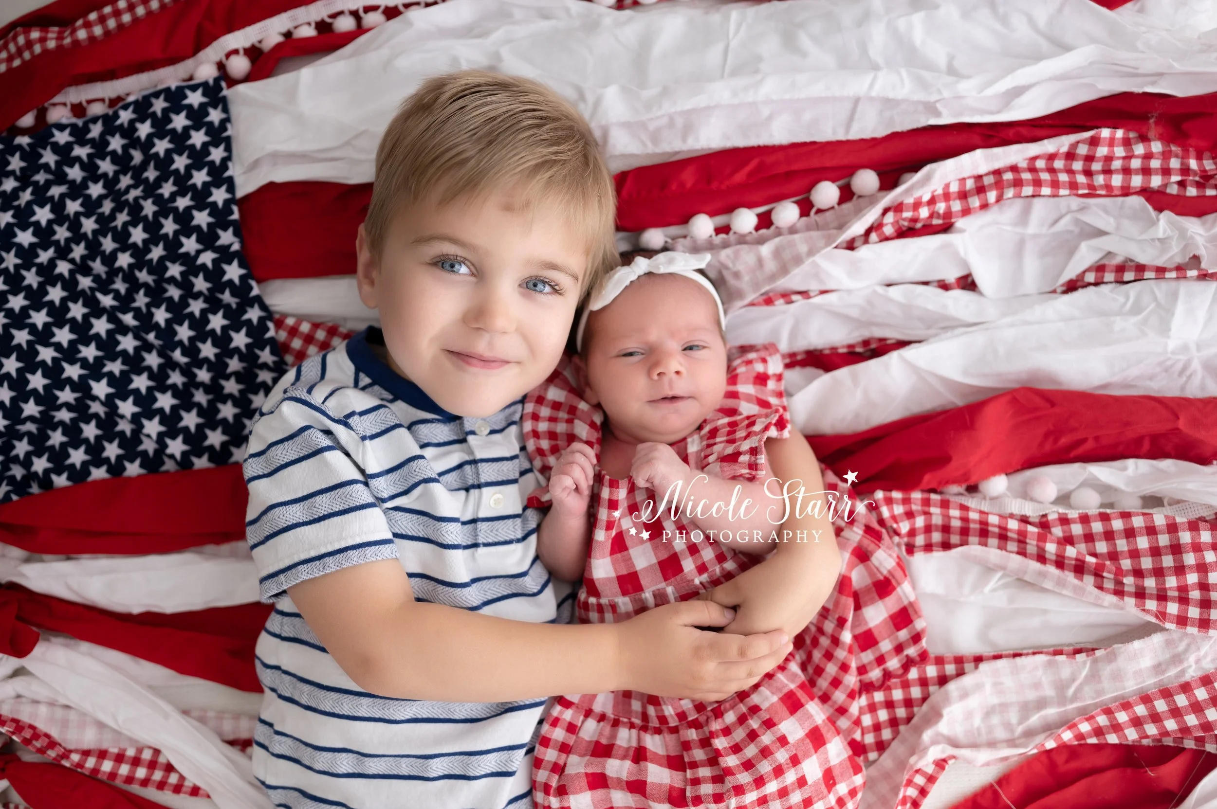 toddler holds baby sister in red gingham dress laying on red and white backdrop during 4th of July portraits in Saratoga Springs studio